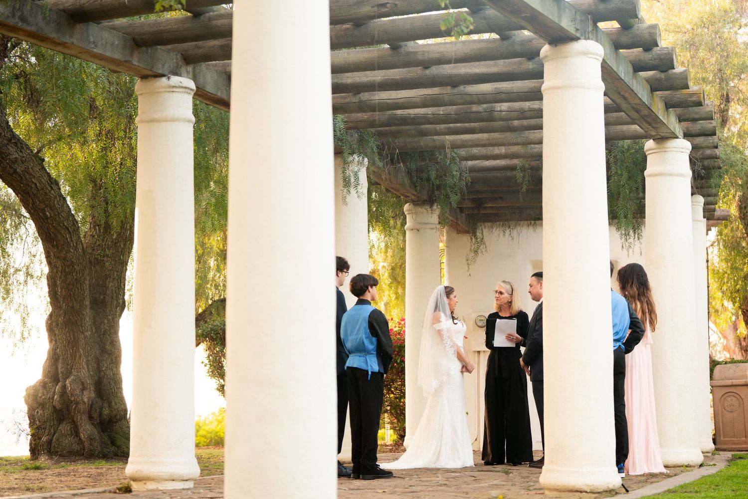 A wedding ceremony takes place under a columned pergola with white pillars and wooden beams in an outdoor garden setting.