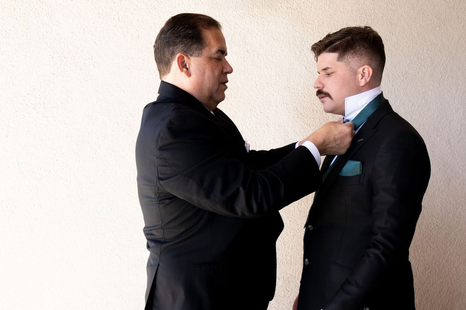 Two men in formal black suits adjusting a bowtie during wedding preparation.