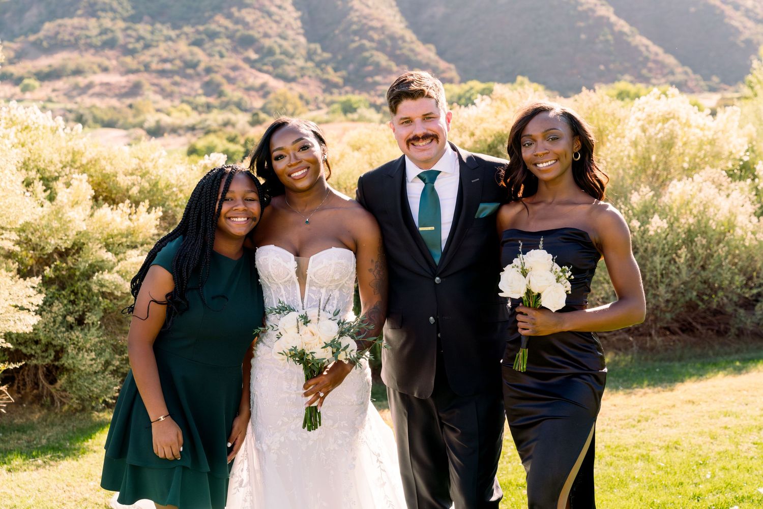 Wedding party poses together outdoors with scenic mountain backdrop on a sunny day.