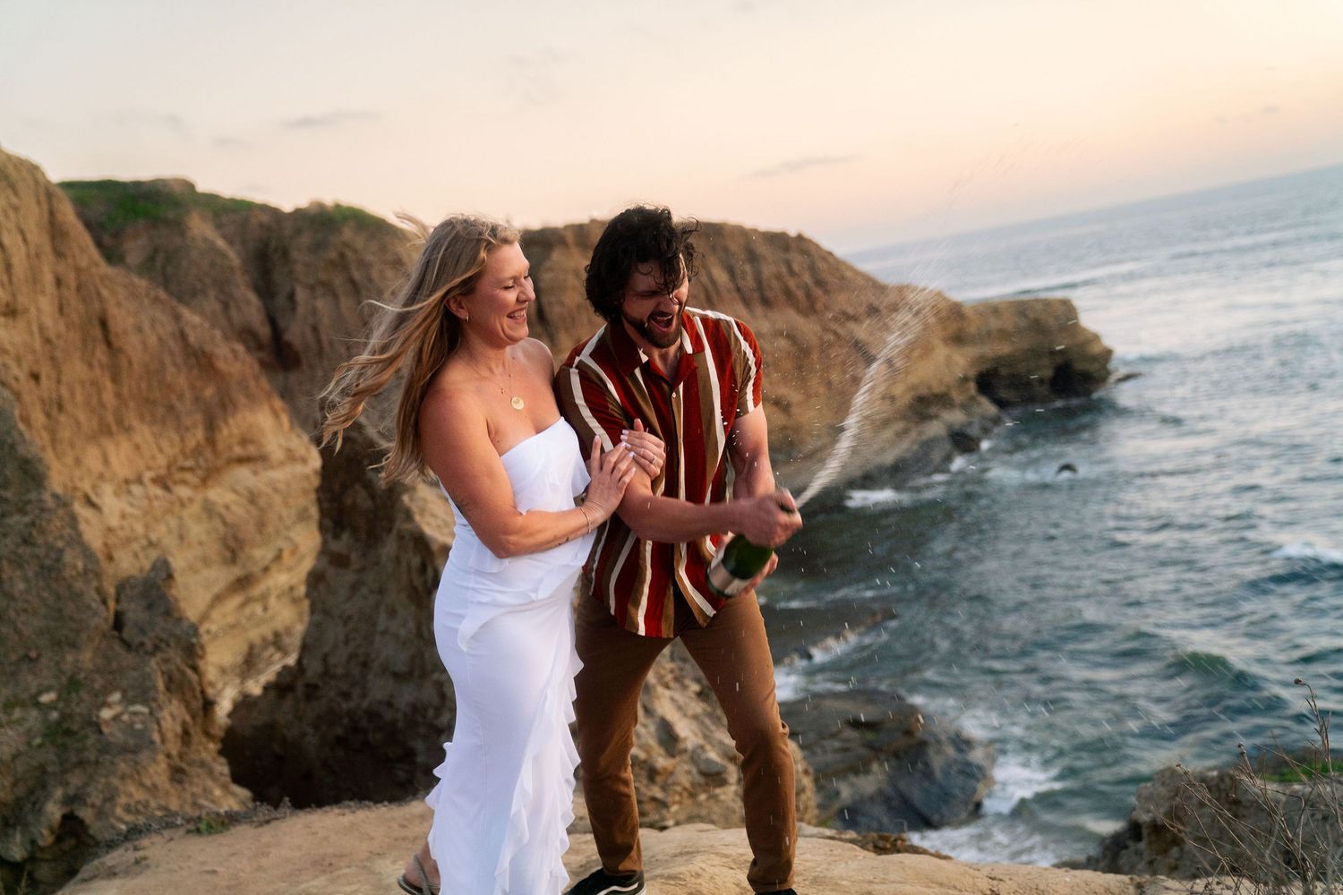 A couple in wedding attire poses at sunset on dramatic coastal cliffs overlooking the ocean.