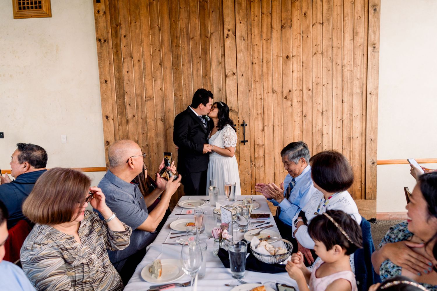 Wedding guests seated at a dinner table applaud as a couple stands in front of rustic wooden doors during a reception.