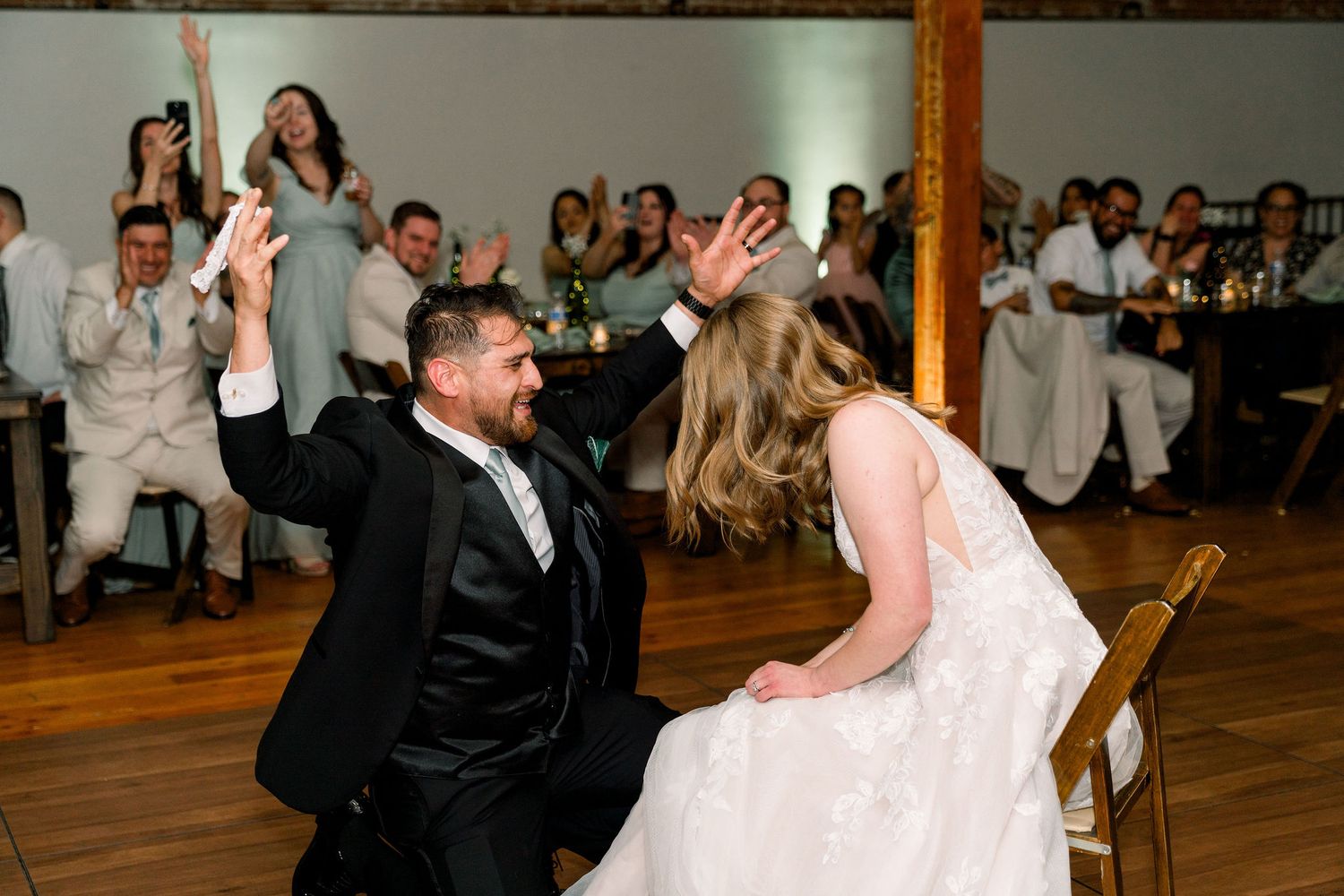 Wedding couple plays a fun game during their reception on a wooden dance floor while guests cheer in the background.