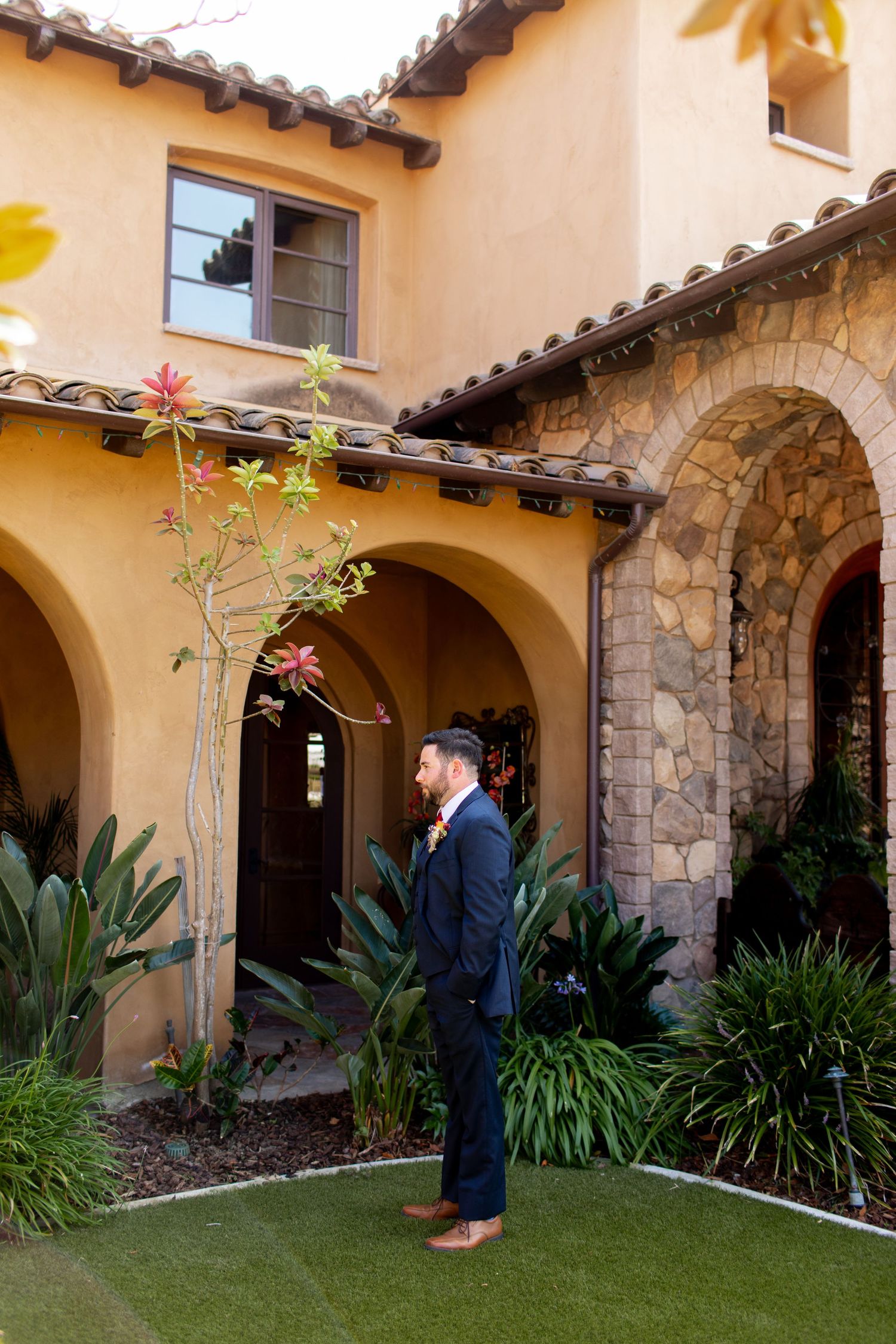 A groom waits in a Mediterranean-style courtyard with stone arches and lush landscaping.