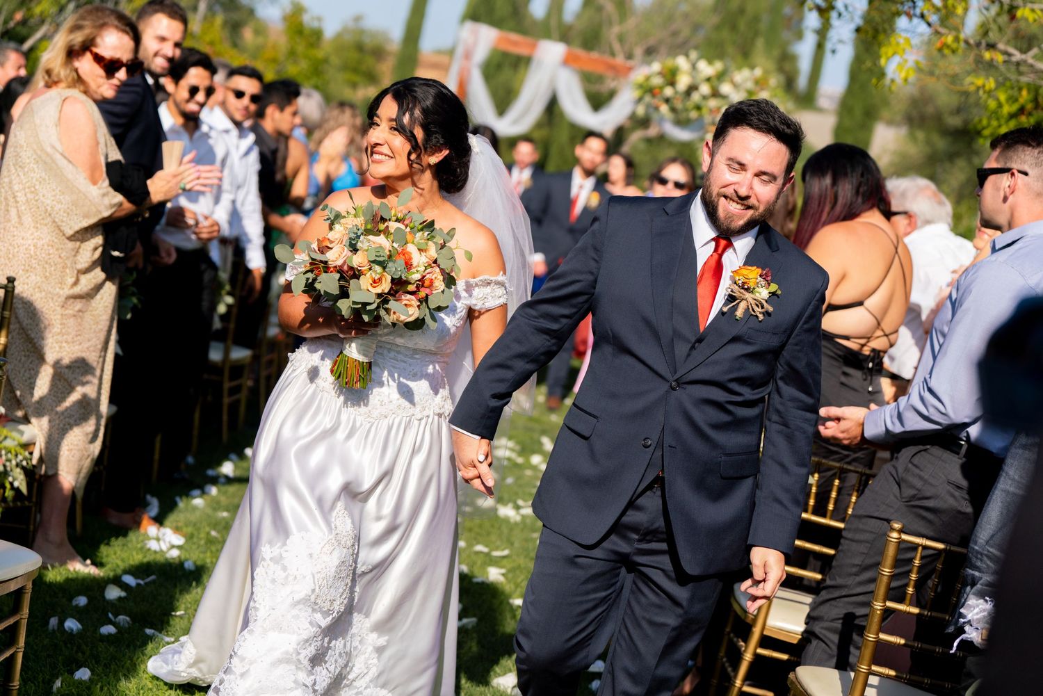 Happy newlyweds exit their outdoor wedding ceremony while guests shower them with flower petals.