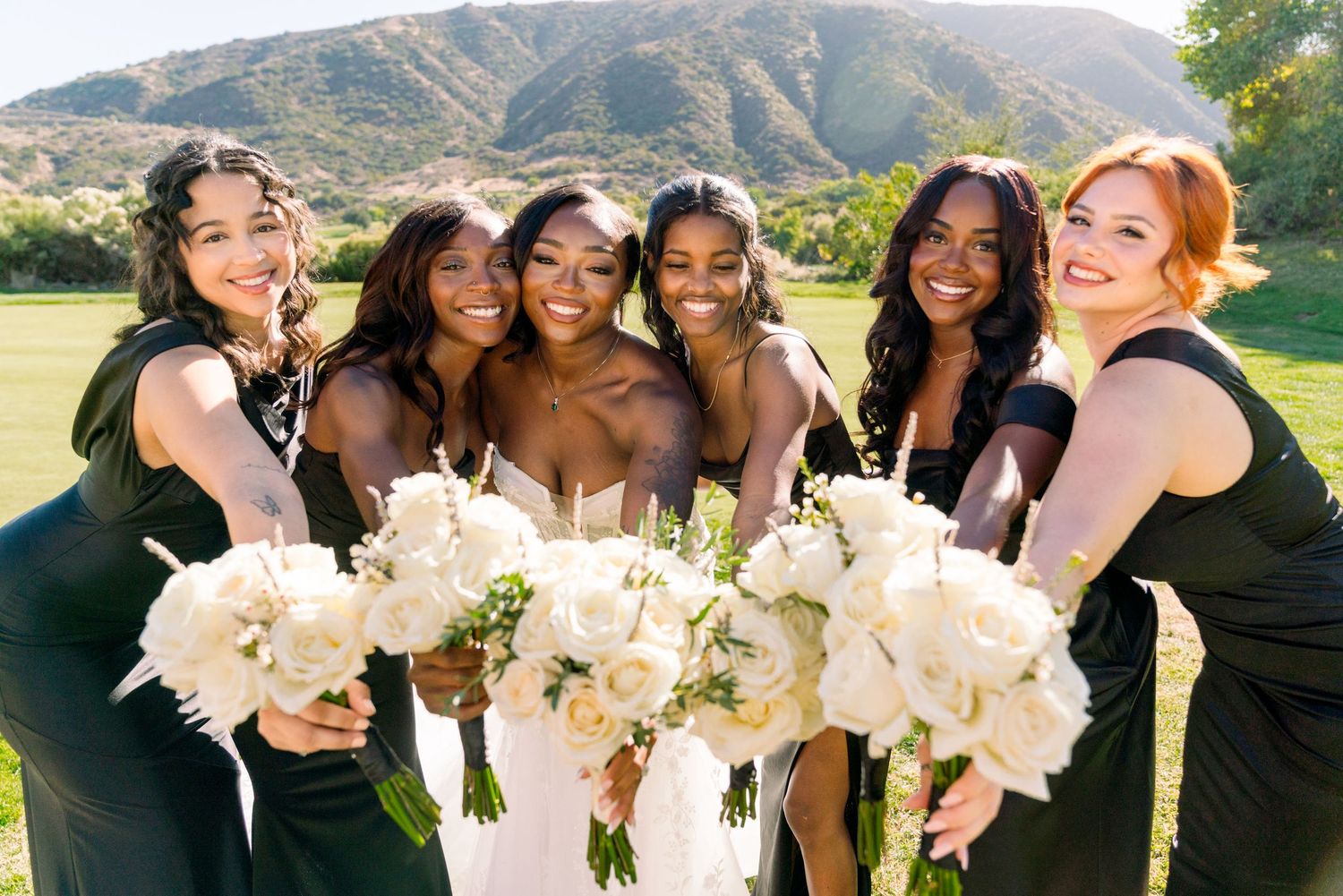 Bridal party in dark dresses holding white bouquets pose together on grass with mountain backdrop.