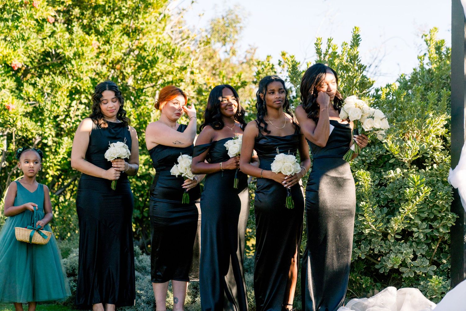 A group of bridesmaids in dark dresses pose together at an outdoor wedding.