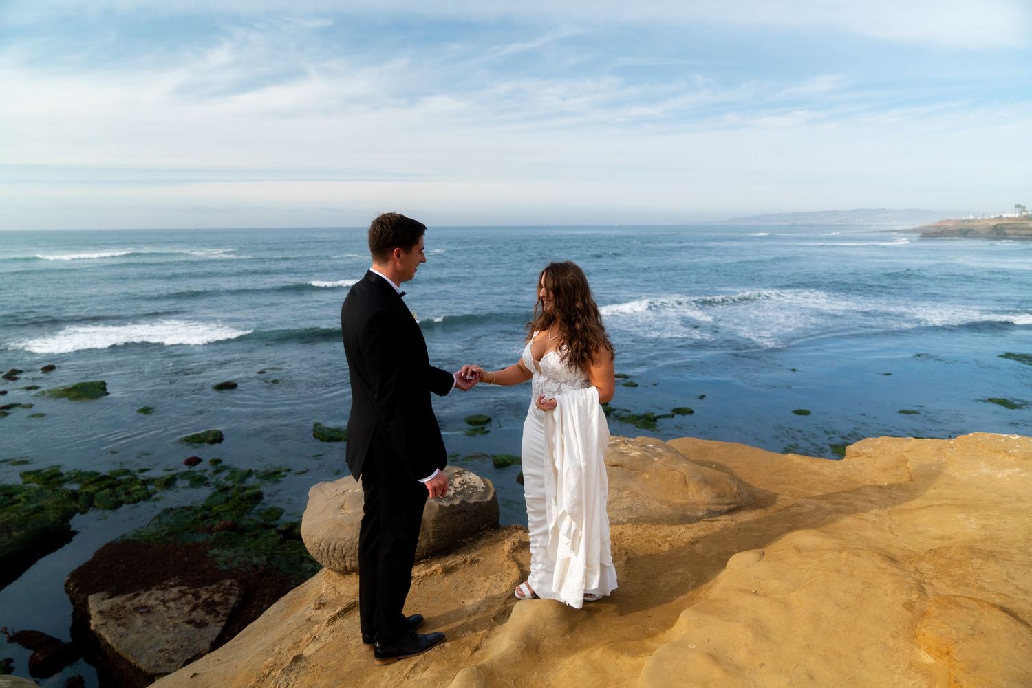 Couple in wedding attire shares a moment on coastal cliffs overlooking the ocean.