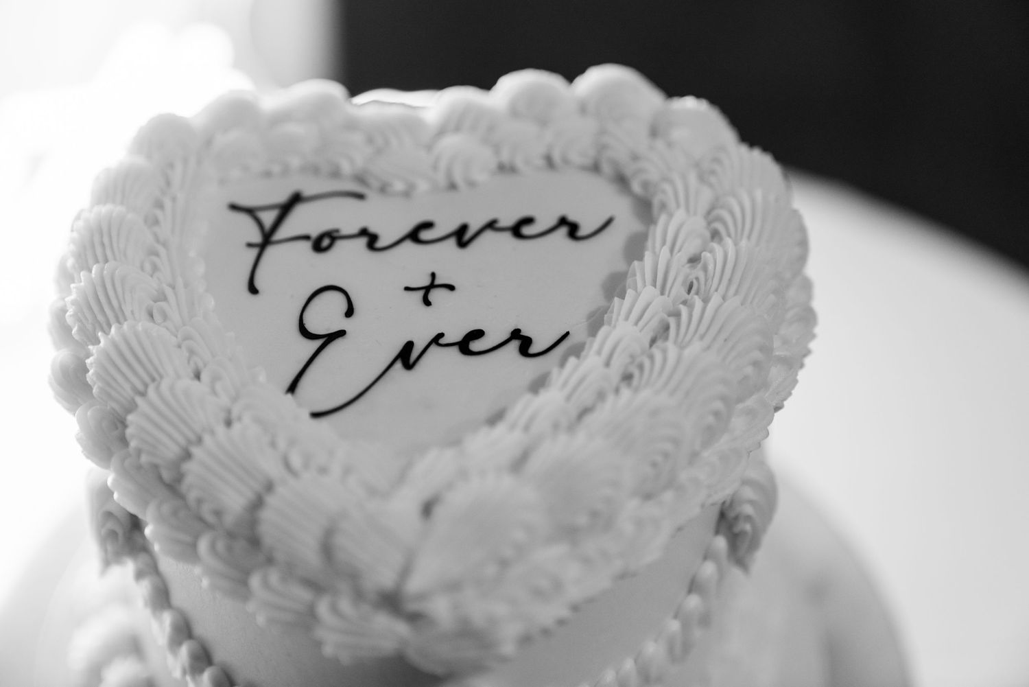 A black and white close-up of a wedding cake with 'Forever & Ever' written in script on a heart-shaped decoration.