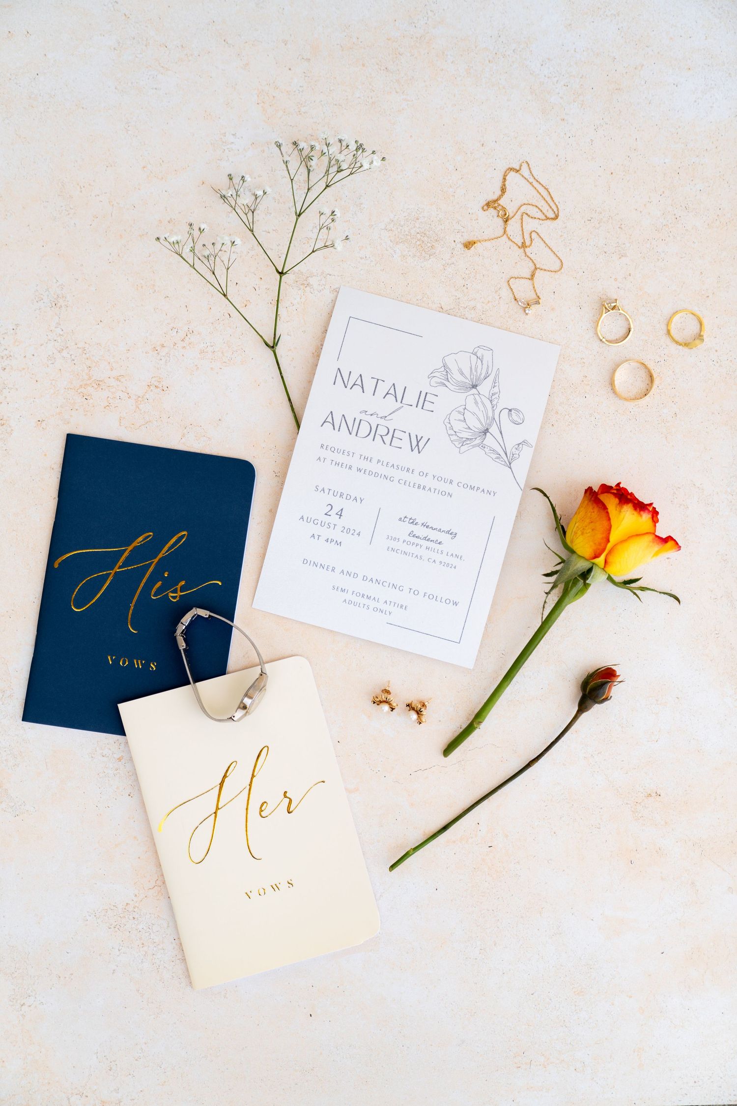 Wedding details flatlay with invitation, vow books, jewelry, and orange rose on cream background.