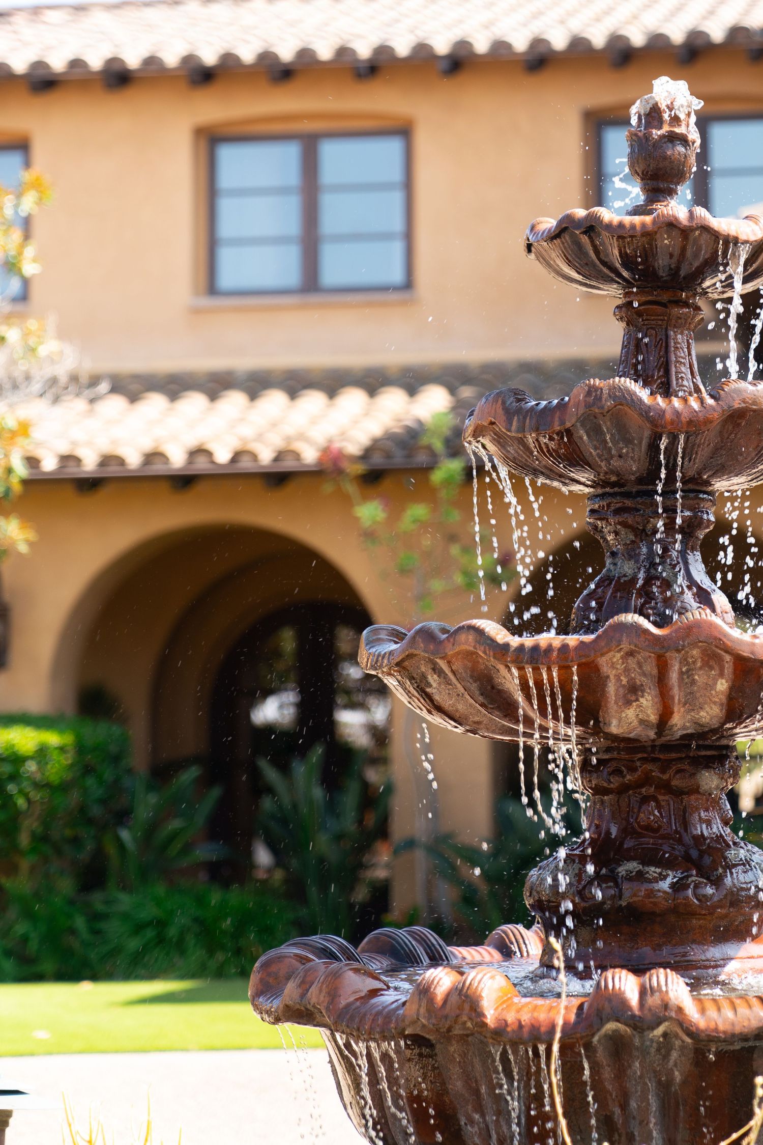Ornate tiered water fountain flowing in front of Spanish-style stucco building with terracotta roof.