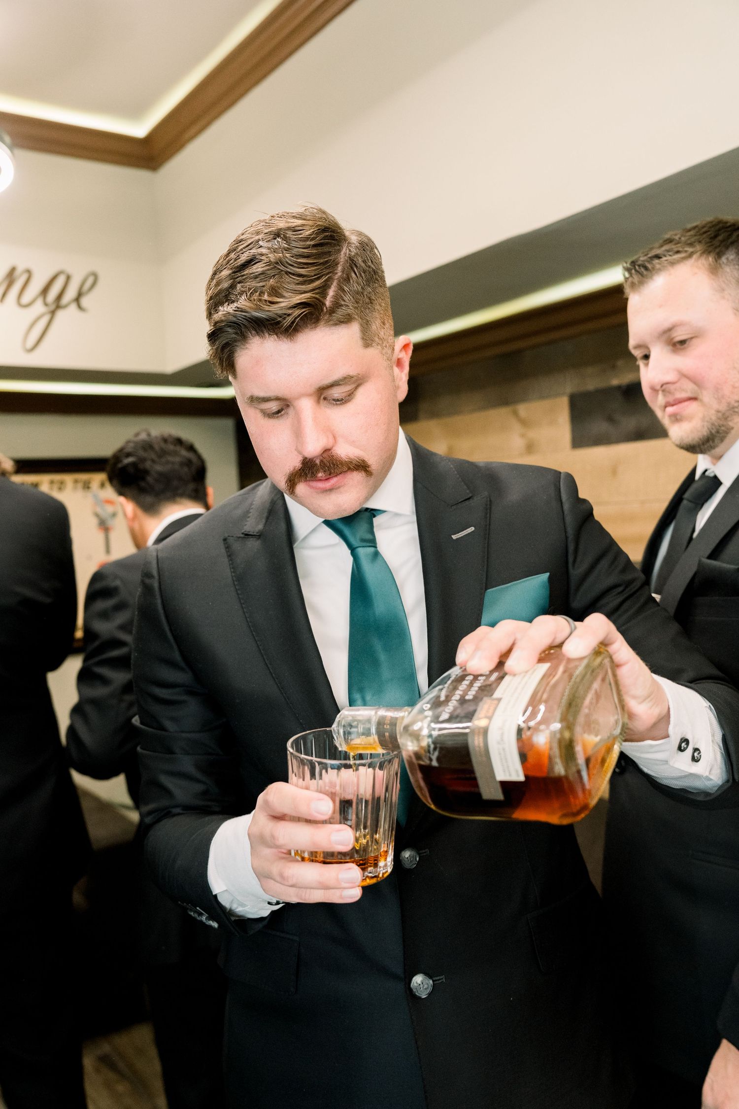 Groomsman in black suit and green tie pours whiskey into glass during wedding reception.