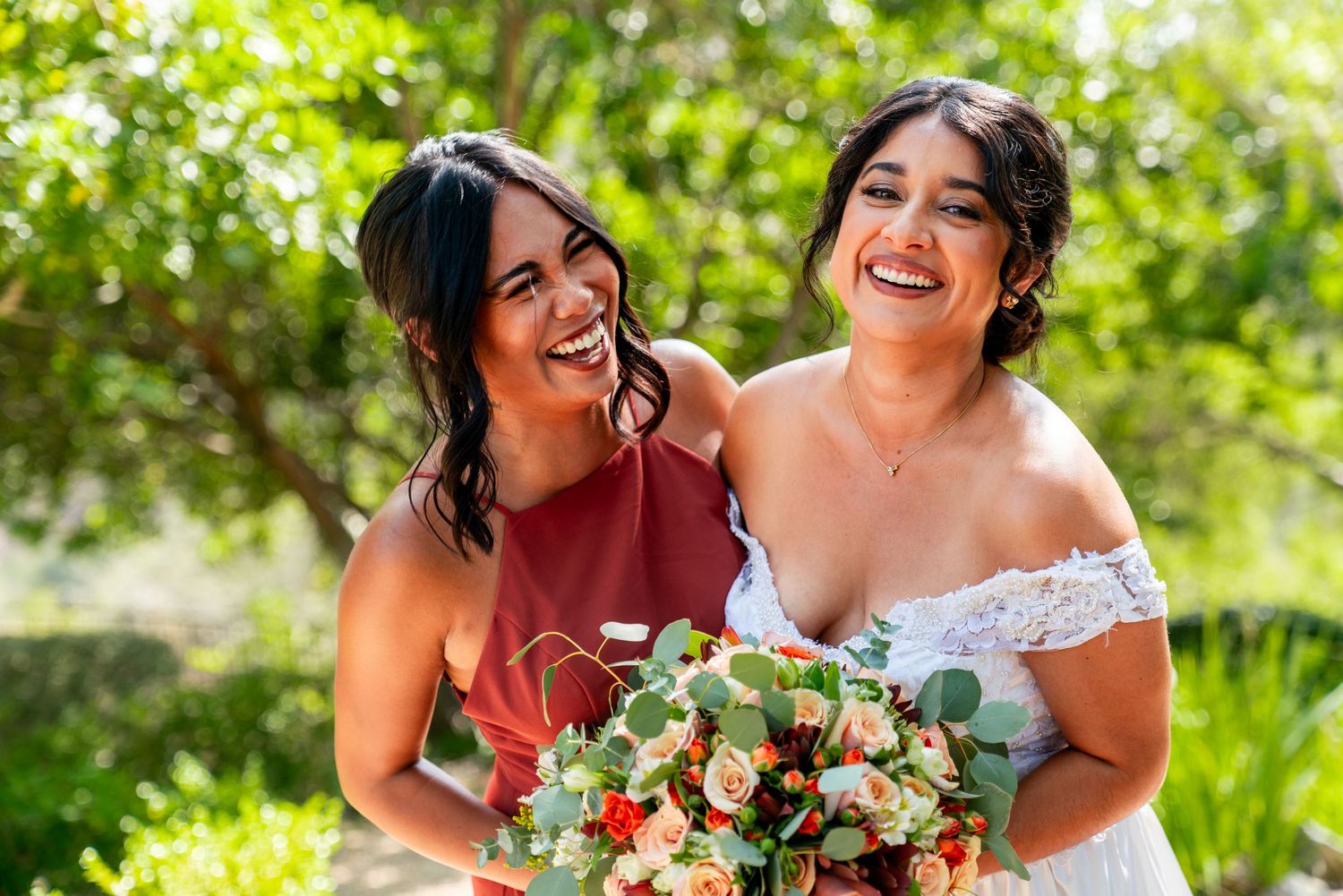Two people in wedding attire share a joyful moment outdoors while holding a beautiful floral bouquet with roses.