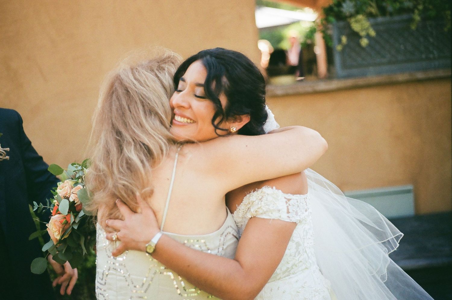 Two women in white dresses share an emotional embrace during a wedding celebration.