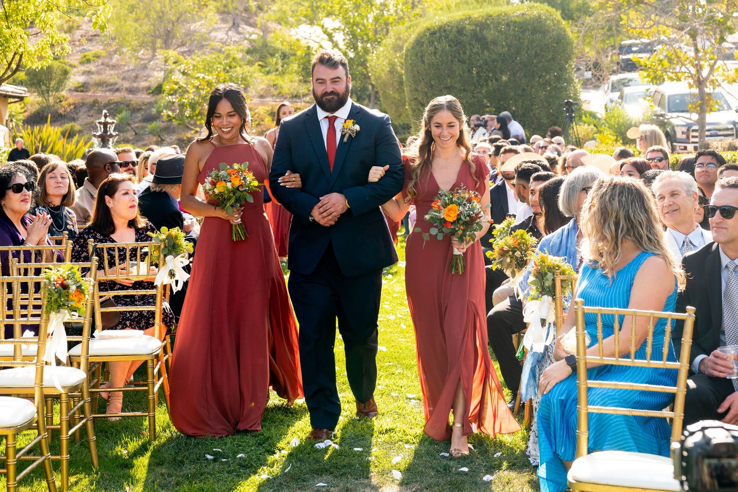 Wedding processional walking down outdoor aisle with burgundy bridesmaids dresses and gold chairs.