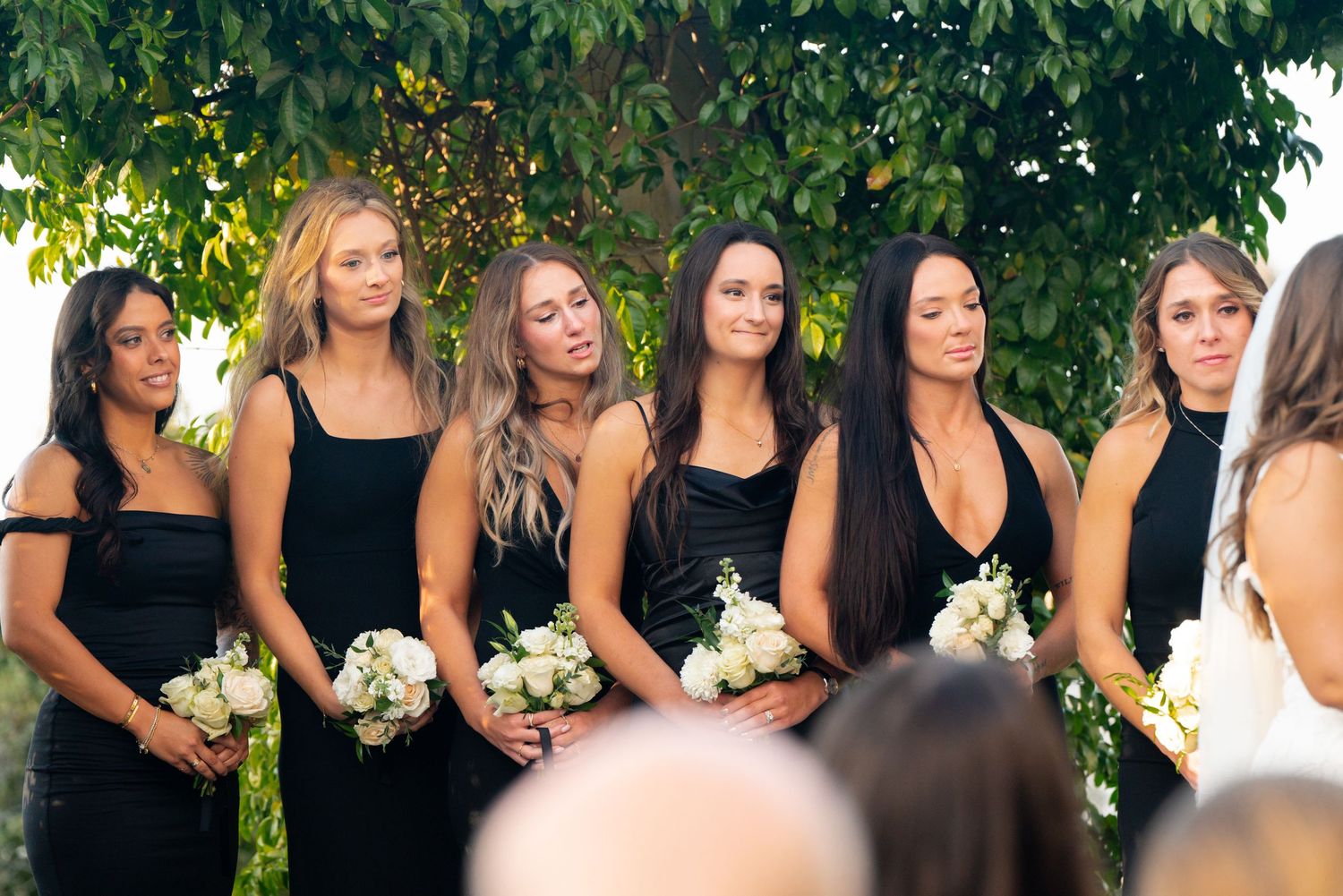 Bridesmaids in matching black dresses hold white bouquets during an outdoor wedding ceremony.