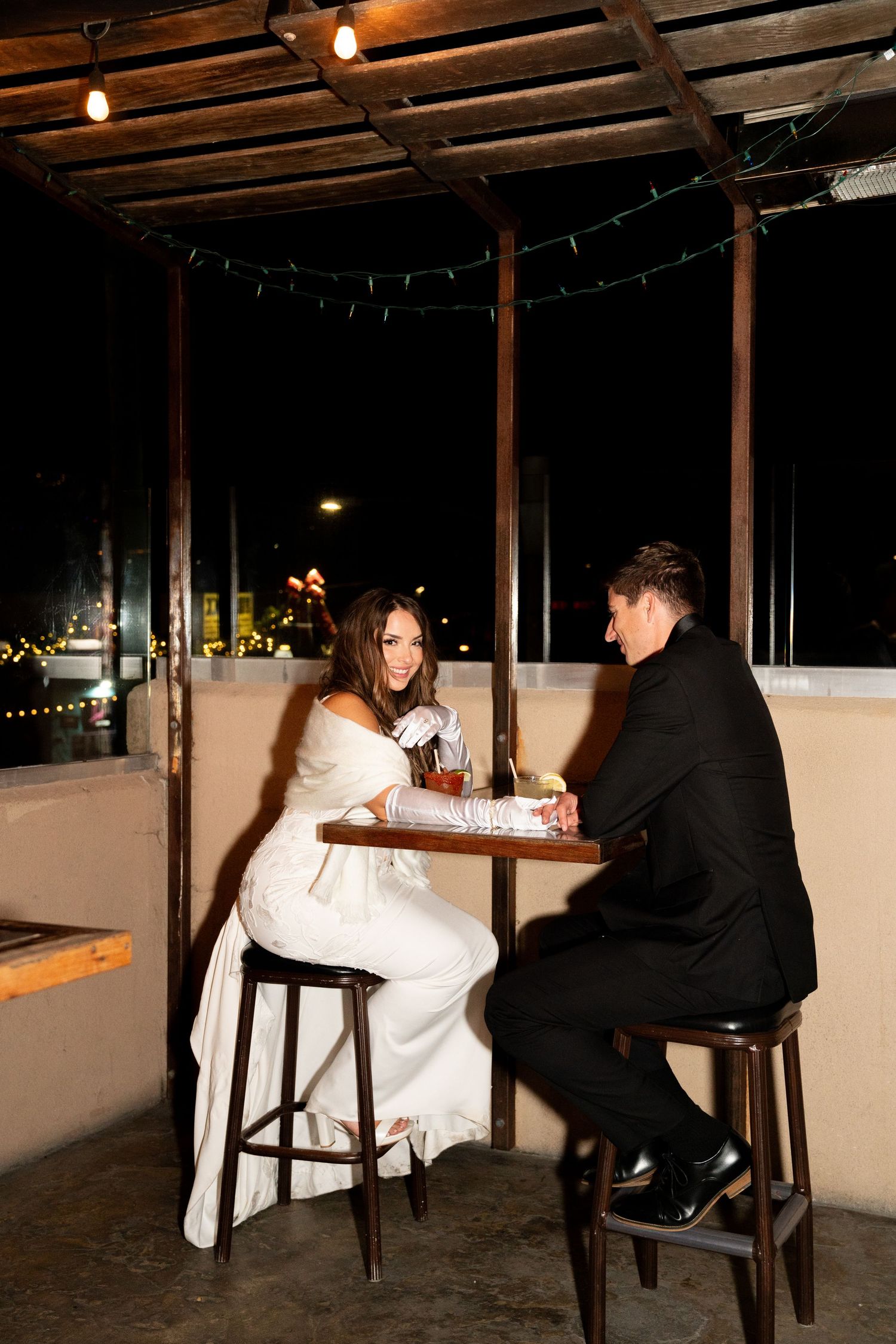 A couple enjoying drinks at a rustic outdoor bar area with string lights overhead during their wedding reception.