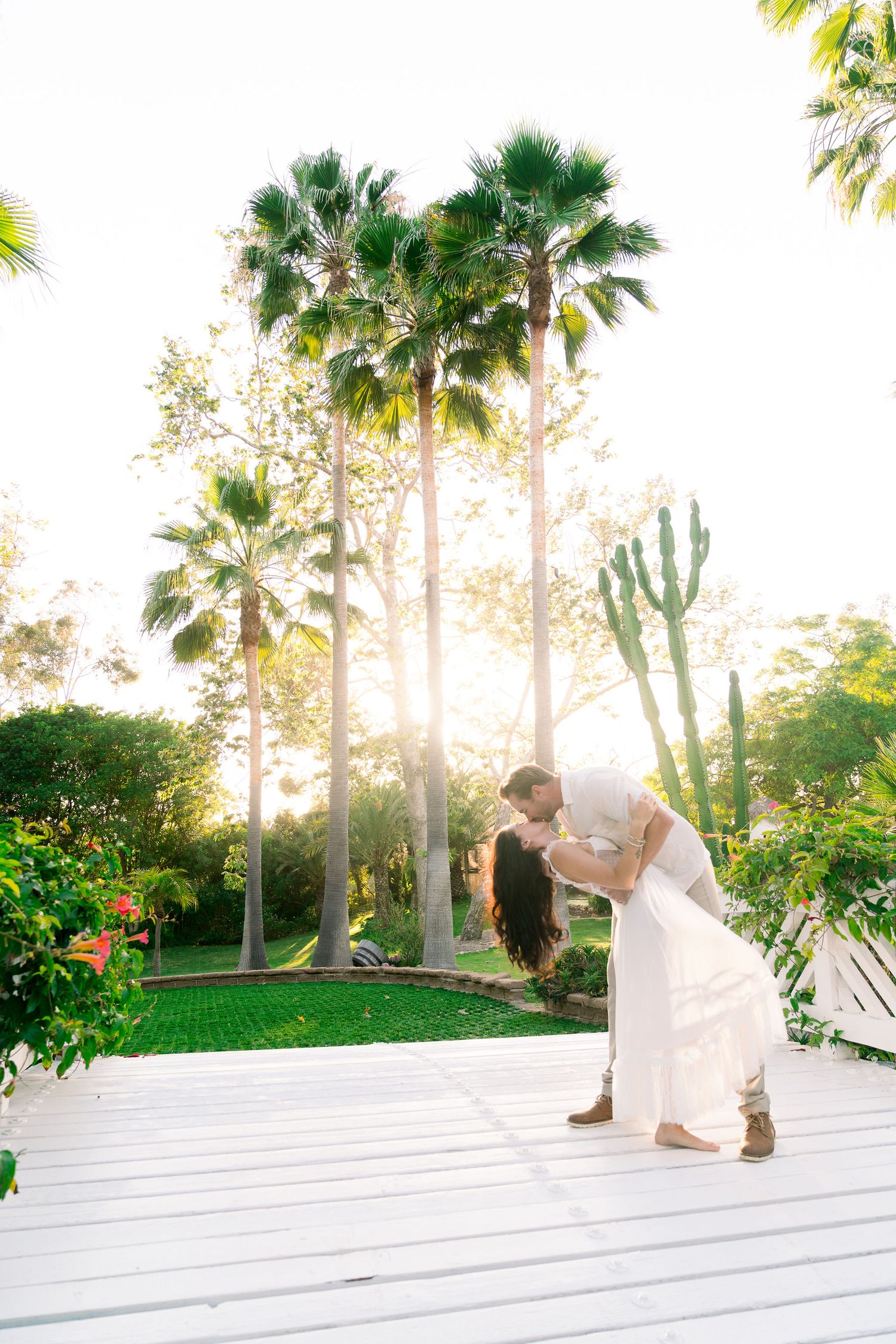A romantic moment captured on a white deck surrounded by palm trees and desert plants in golden sunlight.