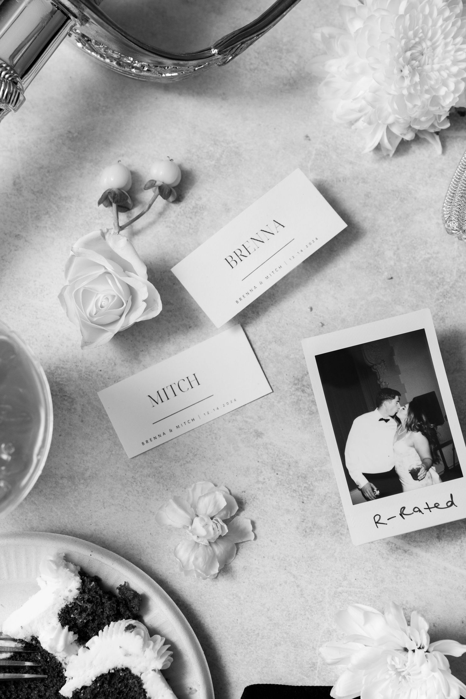 Black and white flatlay with name cards, a Polaroid photo, rose petals, and a plate on a textured surface.