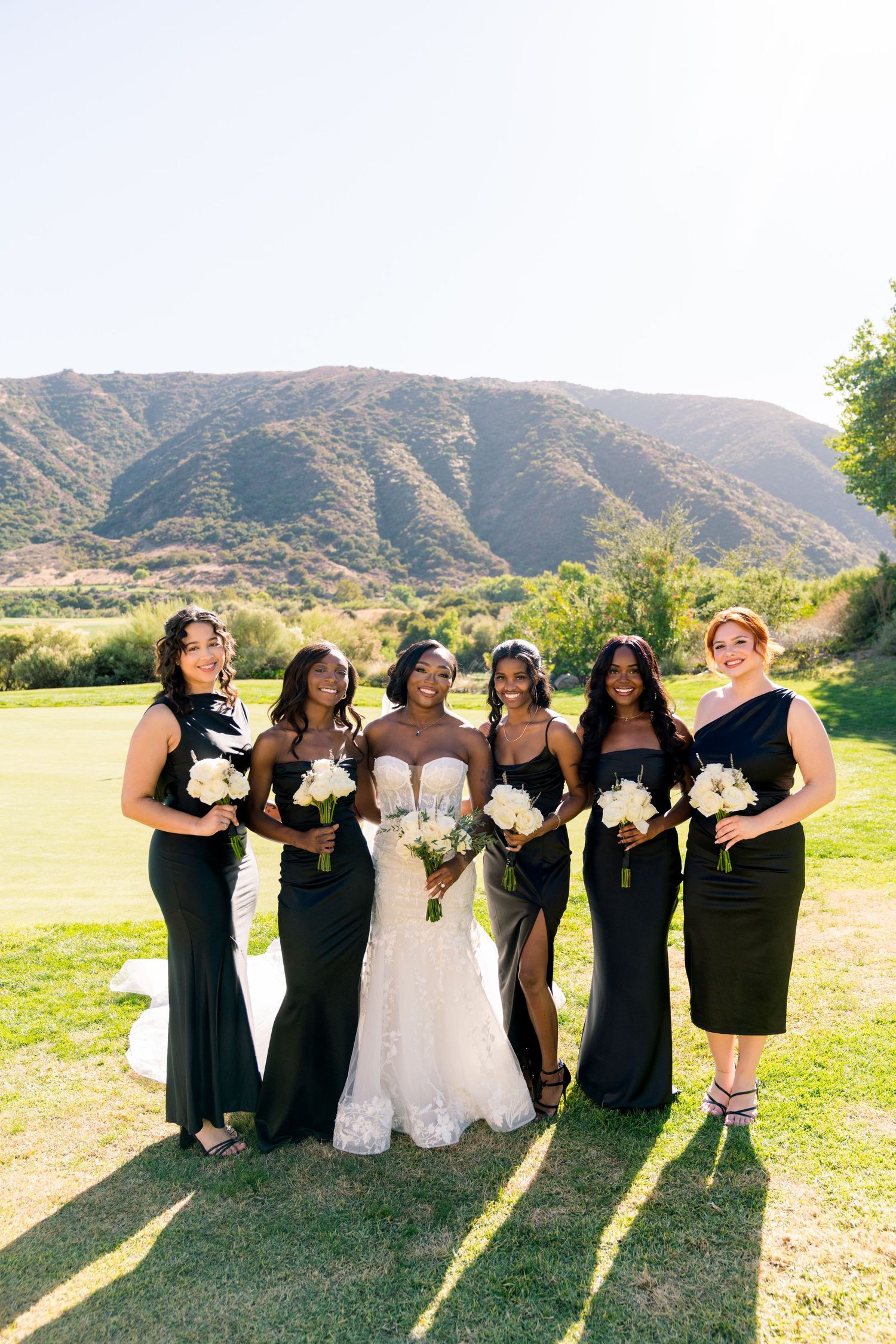 Bridal party wearing black dresses poses with bride in white gown on scenic golf course against mountain backdrop.