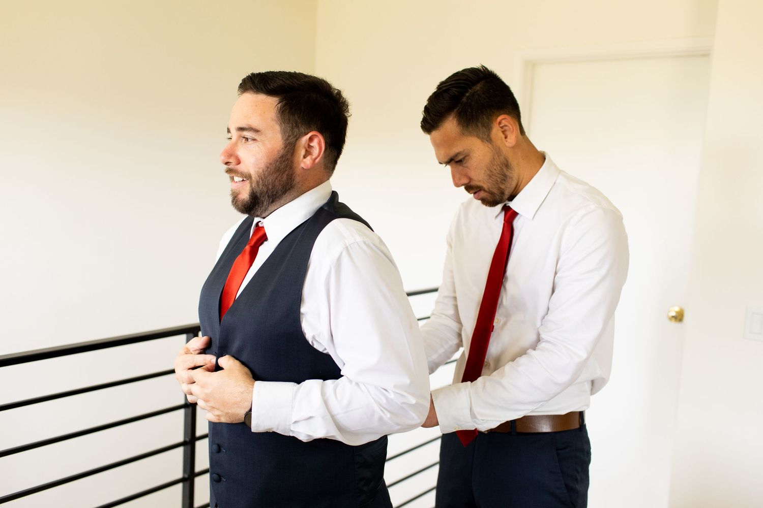 Two people in formal attire with red ties and vests prepare for a special occasion indoors.