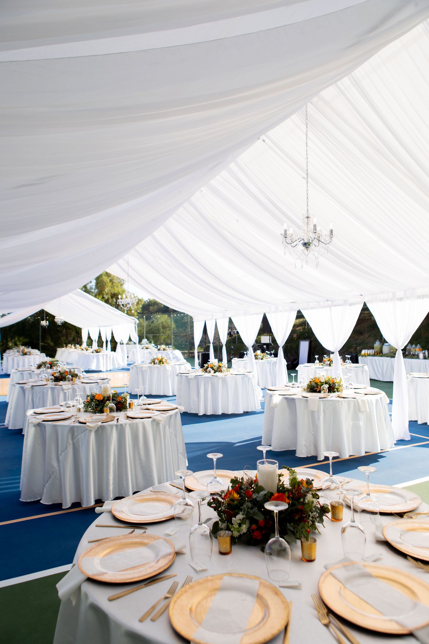 Elegant wedding reception setup with white draped tent ceiling, round tables, and gold charger plates on white linens.