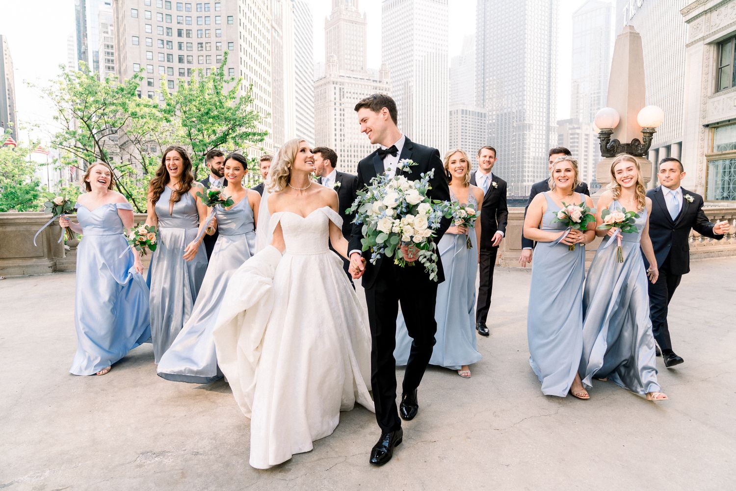 A wedding party in light blue bridesmaid dresses and formal attire walks together on an urban street with tall buildings.