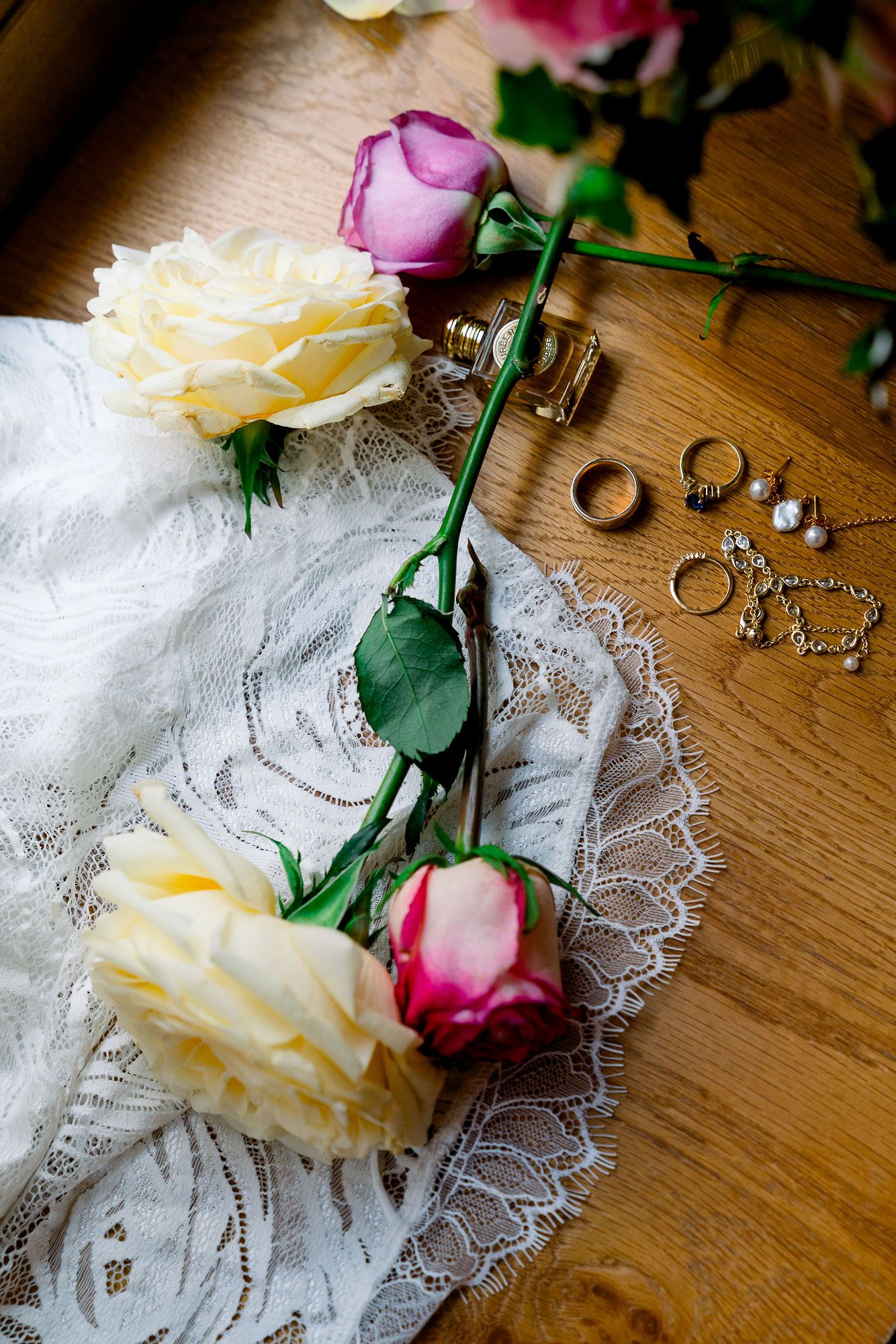 Wedding rings, jewelry and roses with pink and cream petals rest on delicate white lace against a wooden surface.
