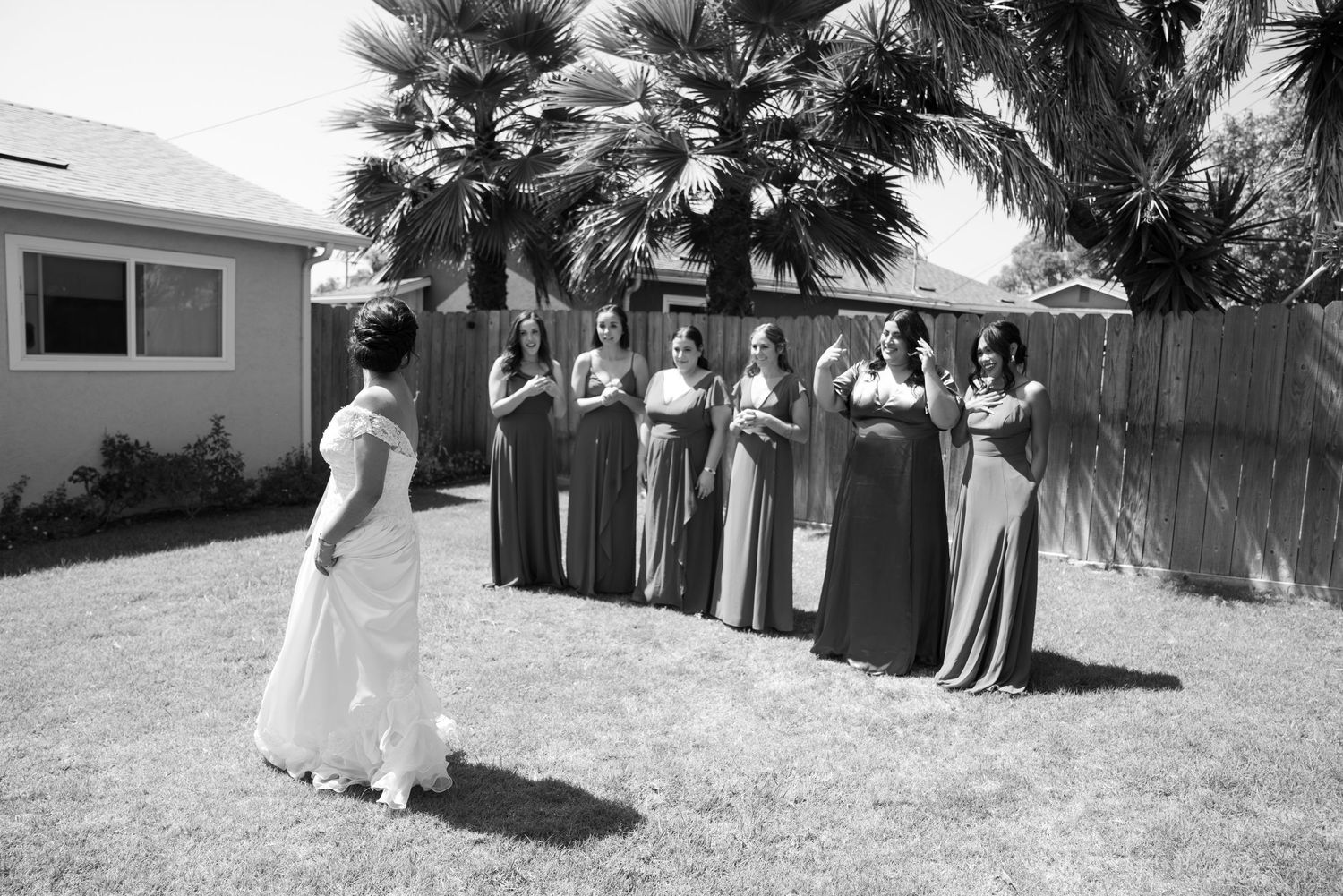 Bridesmaids in long dark dresses stand in line on a lawn with palm trees during a wedding.