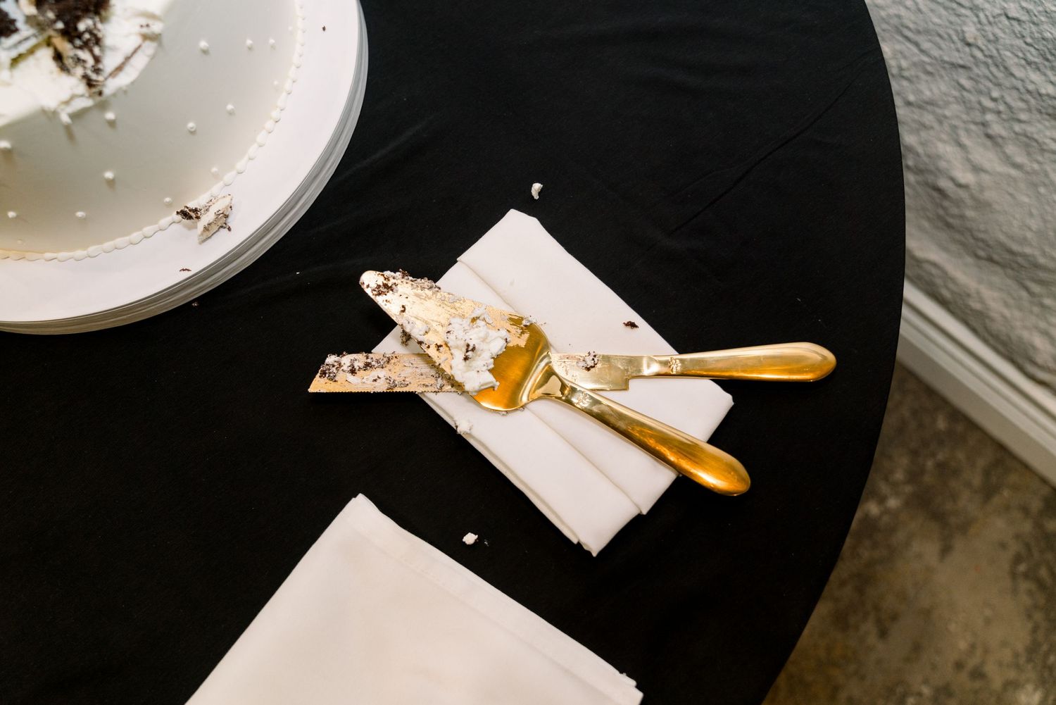 Wedding place setting with white napkin and gold utensils on black tablecloth.