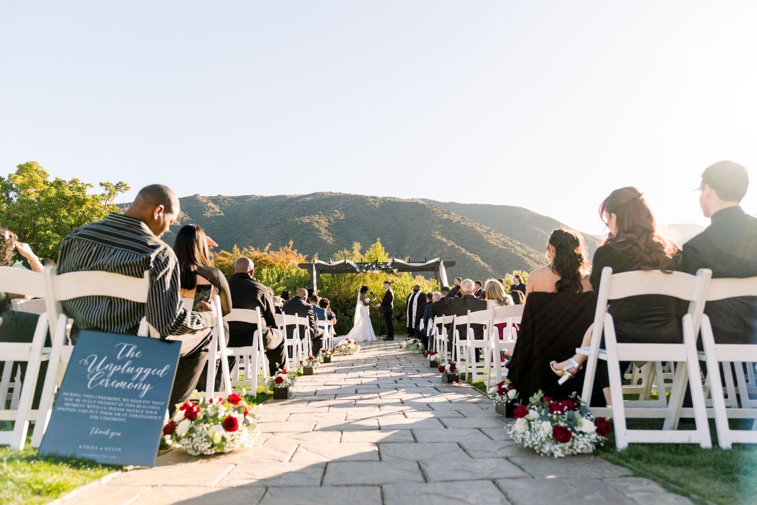 White chairs line a stone pathway for an outdoor wedding with mountains and a welcome sign.