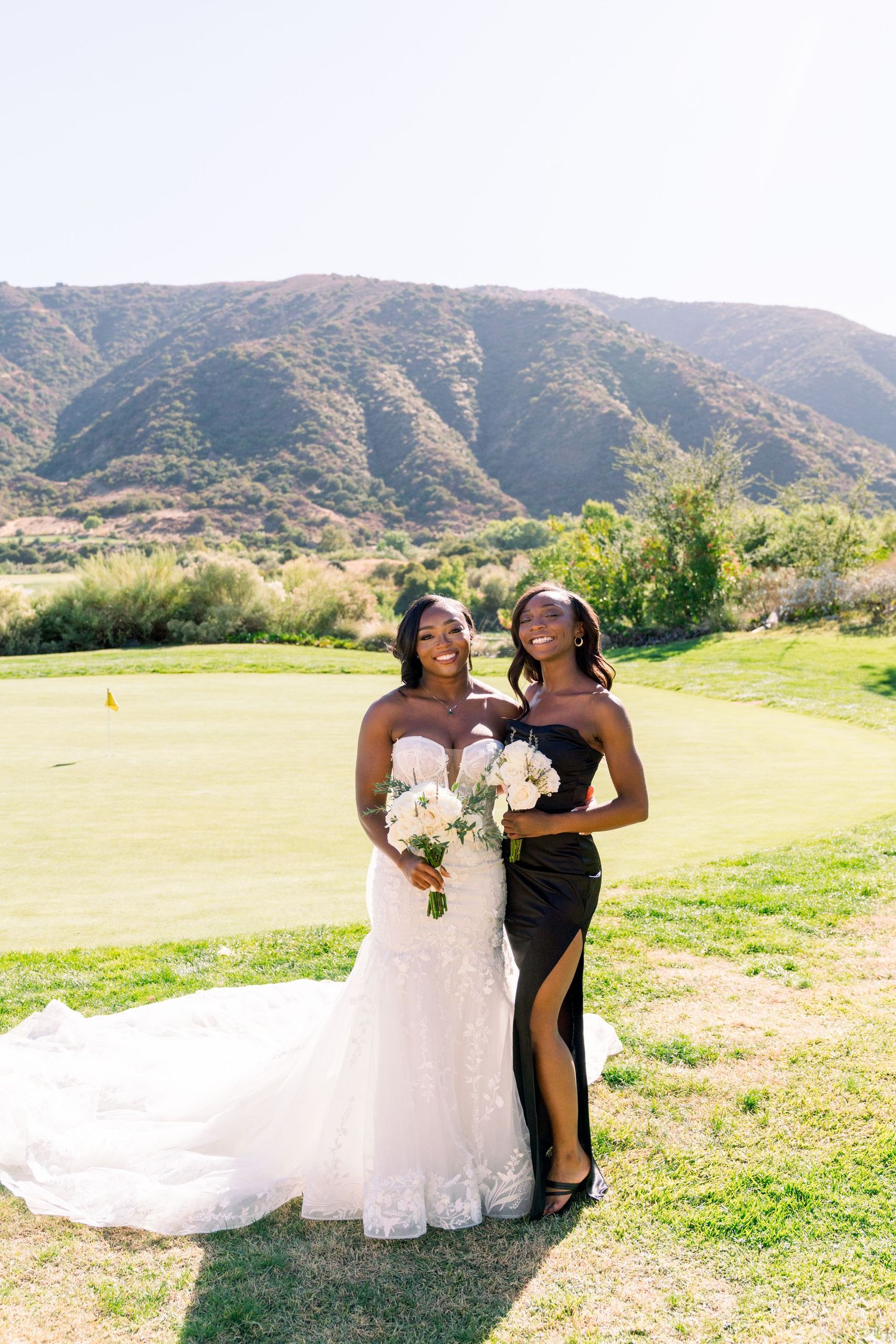 Two bridesmaids pose together on a scenic golf course with mountains in the background at an outdoor wedding.