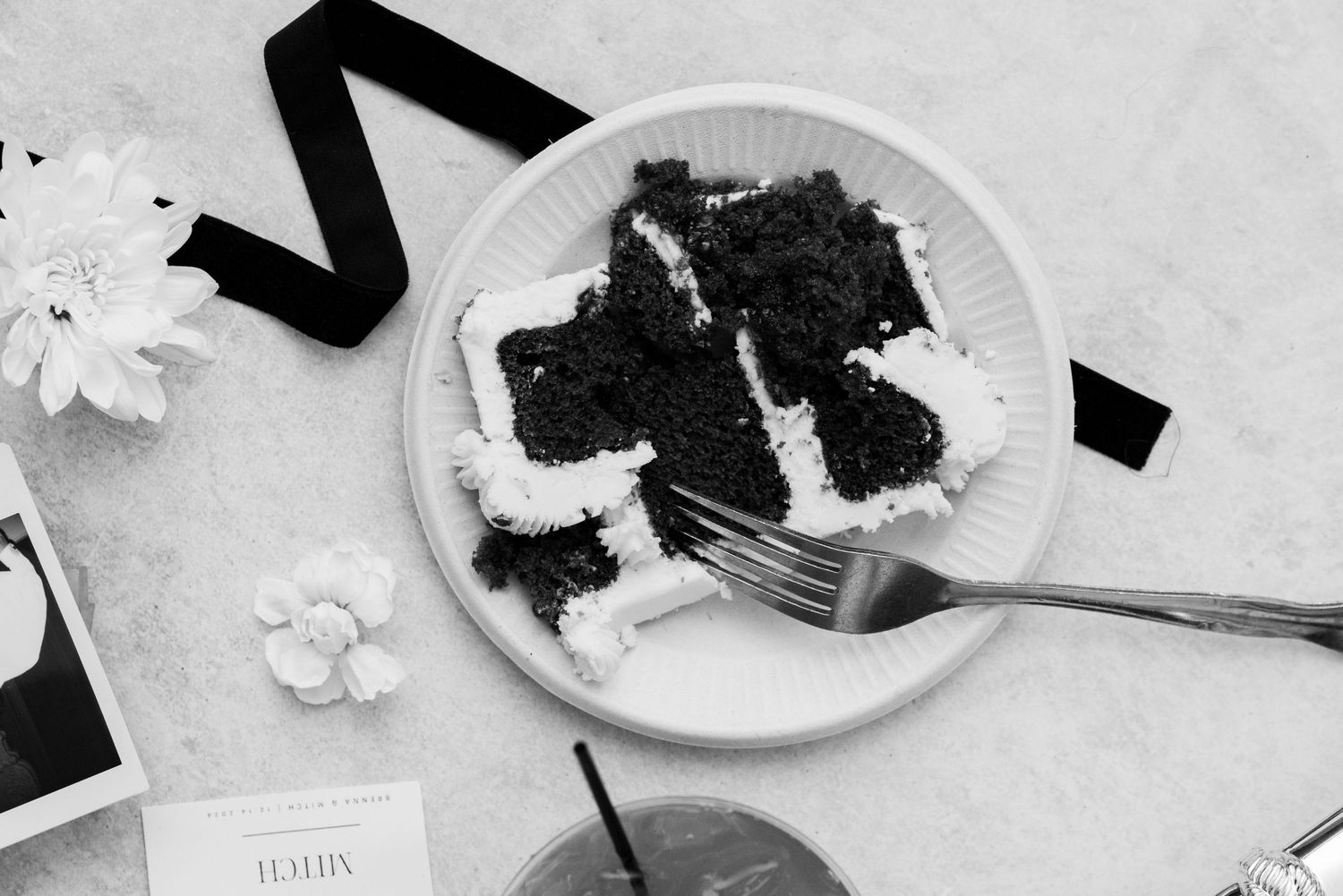 A slice of dark chocolate cake with frosting on a white plate beside black ribbon and white flowers.