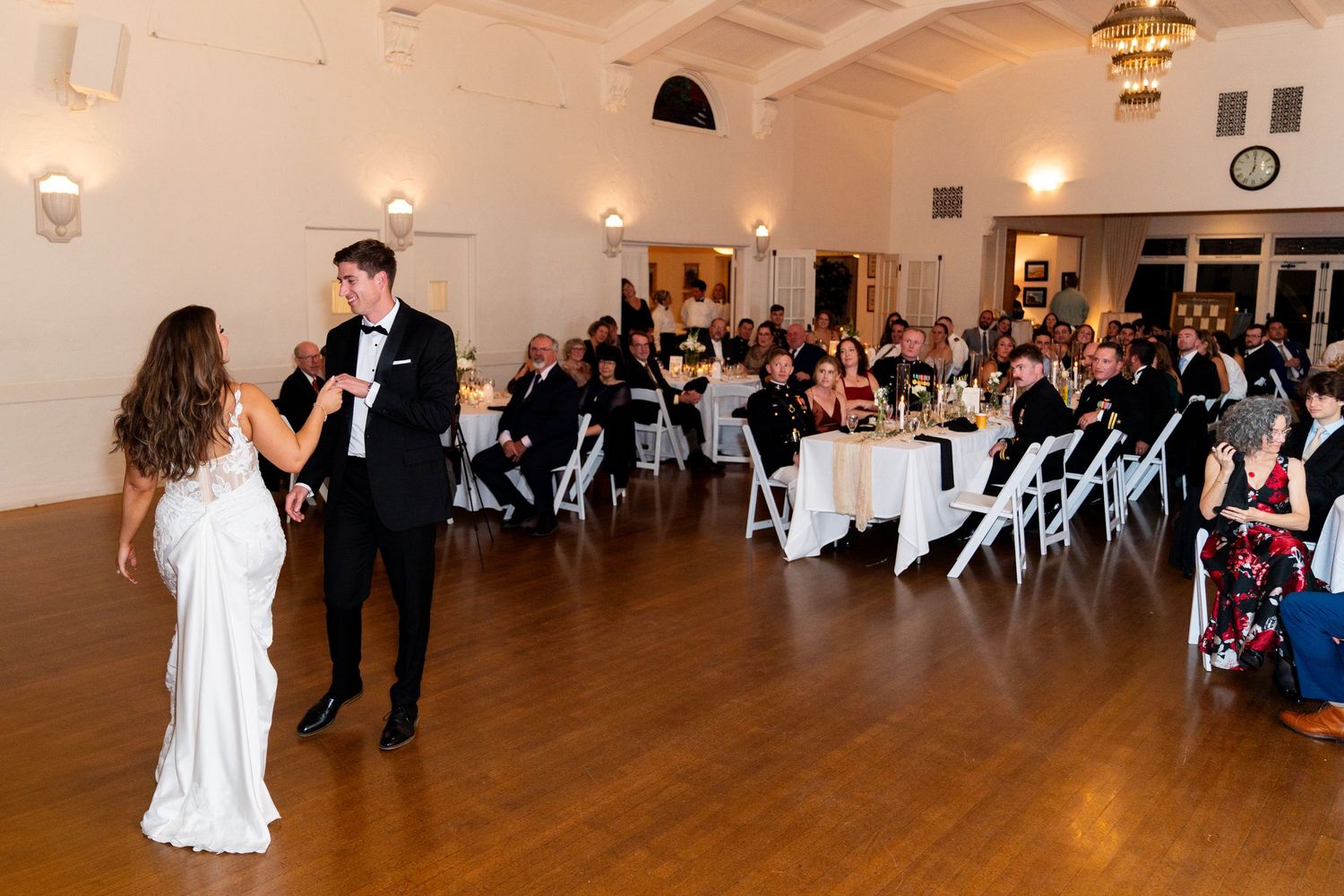 A bride and groom share their first dance in a beautifully lit reception hall with guests watching.