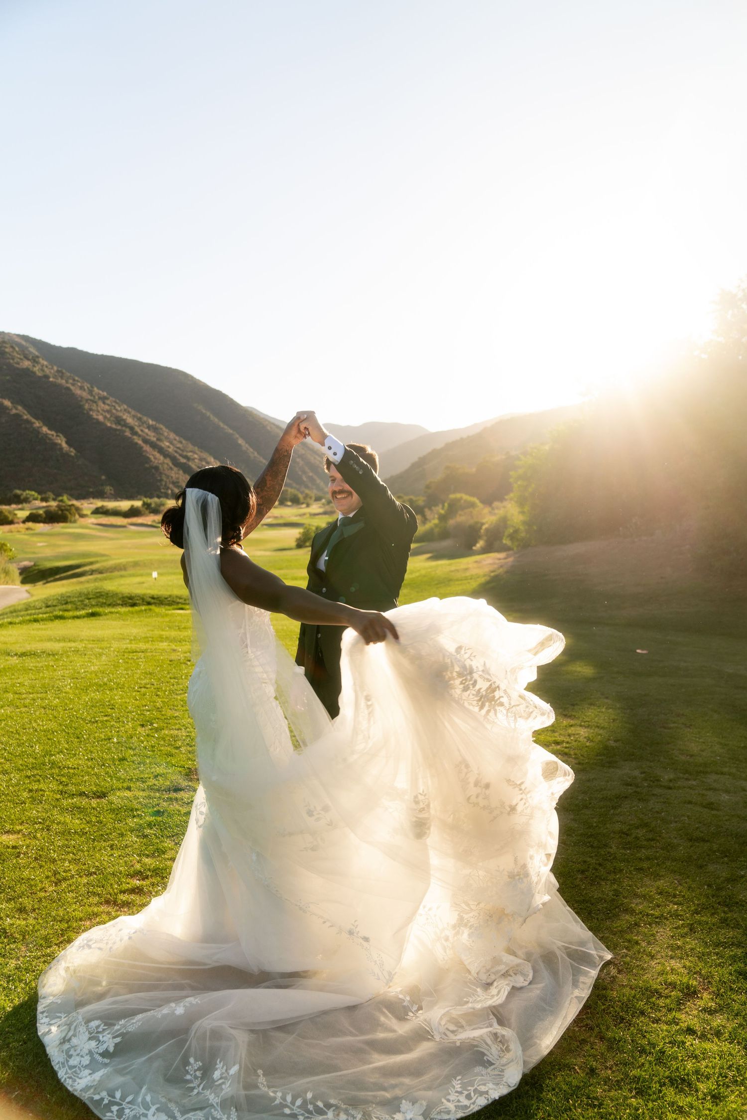 Joyful wedding couple dancing on a golf course during golden hour with mountains in background.
