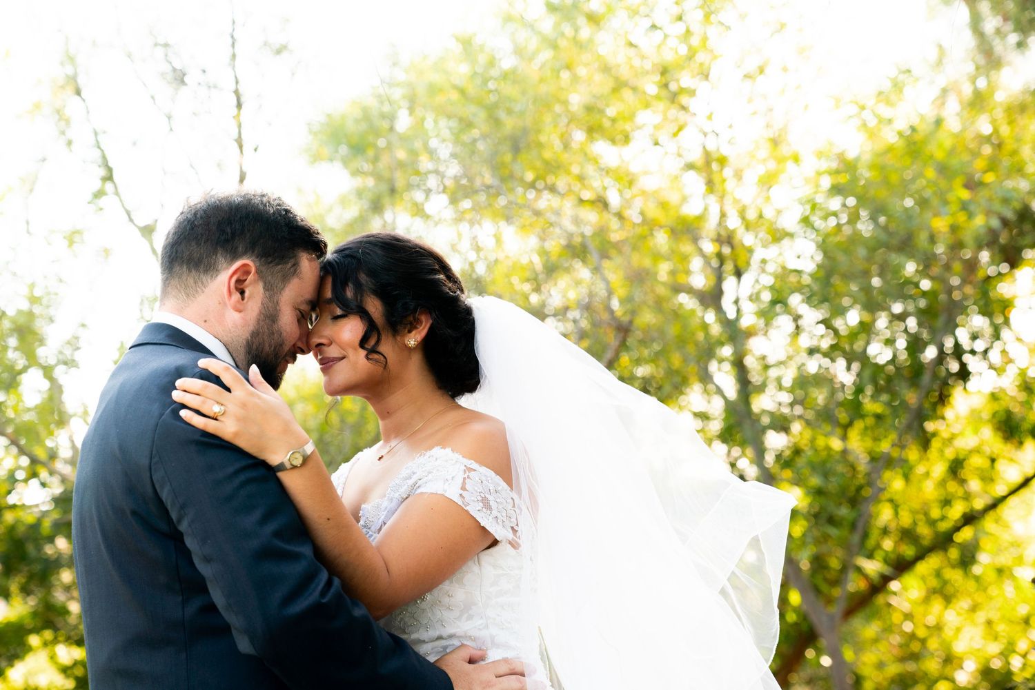 A romantic wedding couple embraces outdoors in a sunlit garden setting with golden foliage in the background.