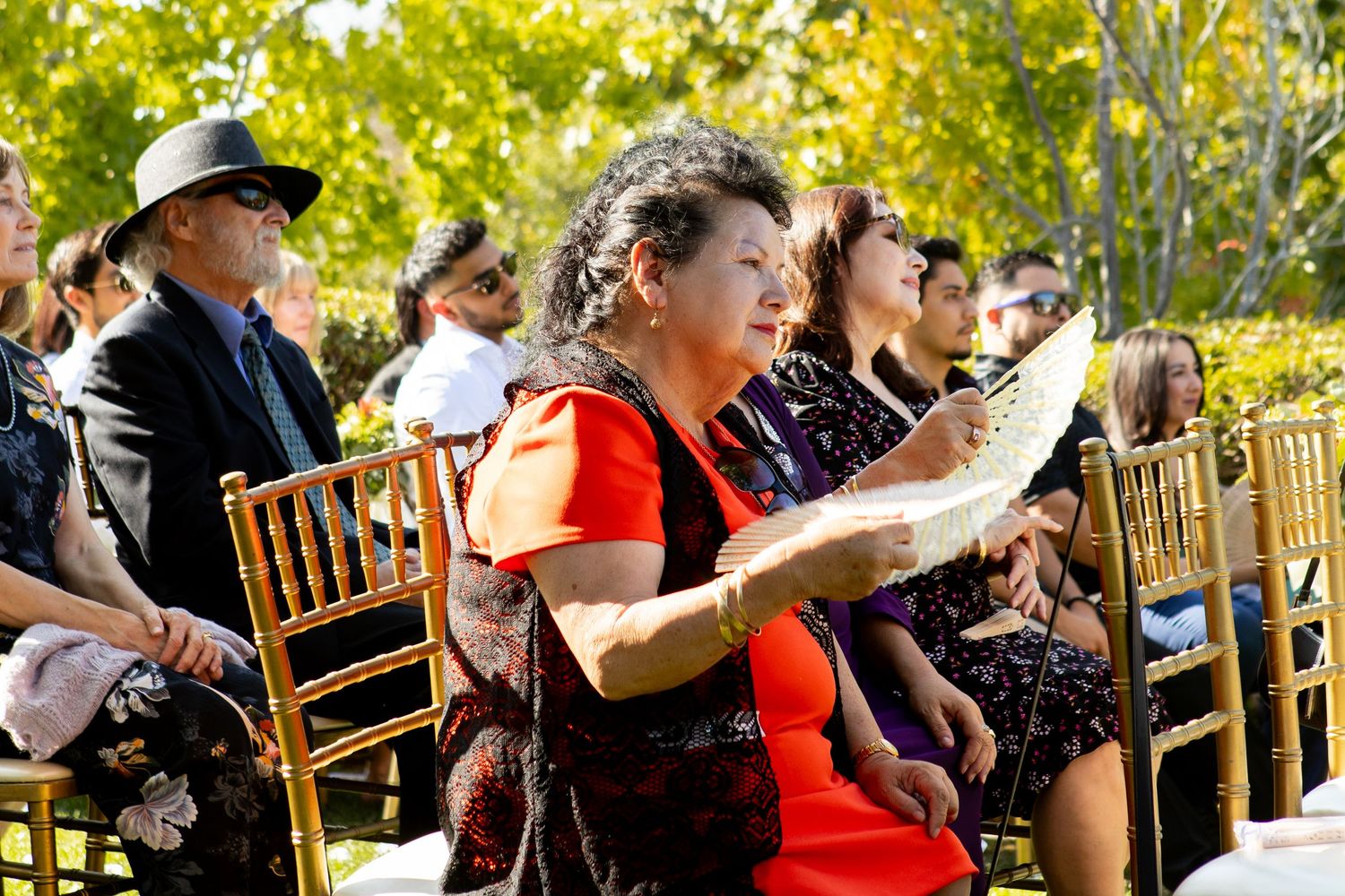 Wedding guests seated in wooden chairs watch ceremony while holding programs in an outdoor garden.