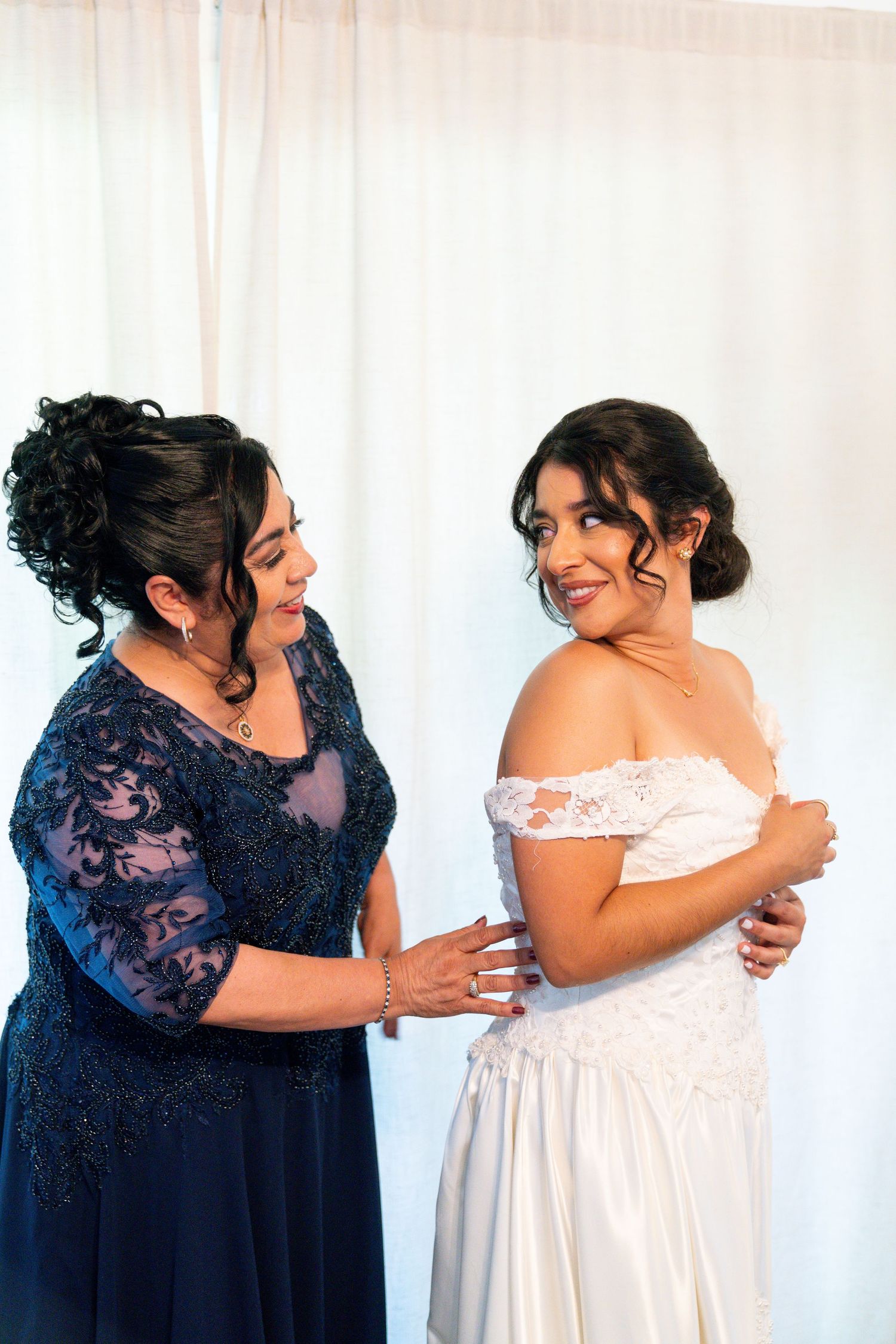 Two women share an intimate moment before a wedding, with one wearing a navy lace gown and the other in white bridal dress.
