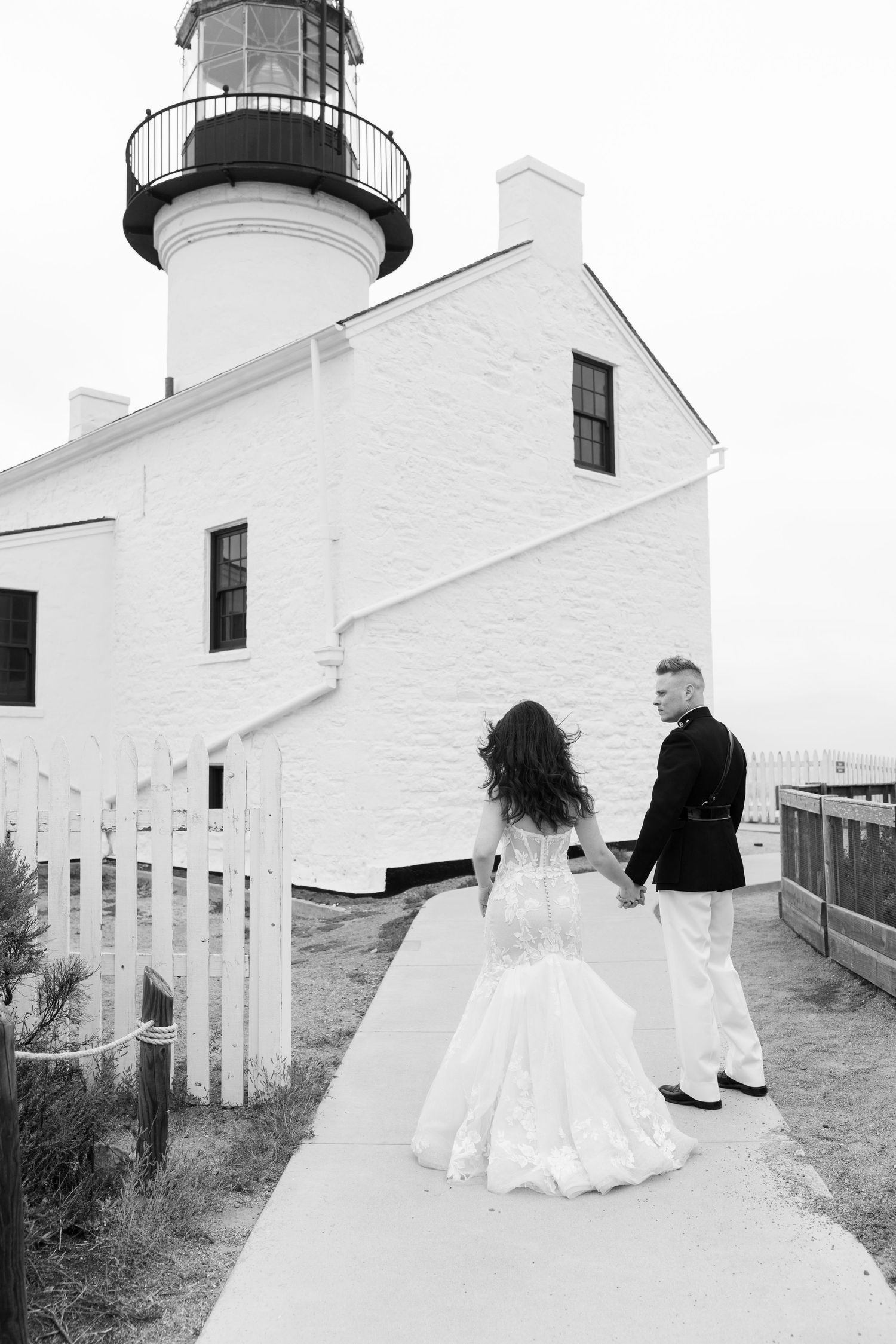Black and white photo of a bride and groom walking toward a white lighthouse with white picket fence.