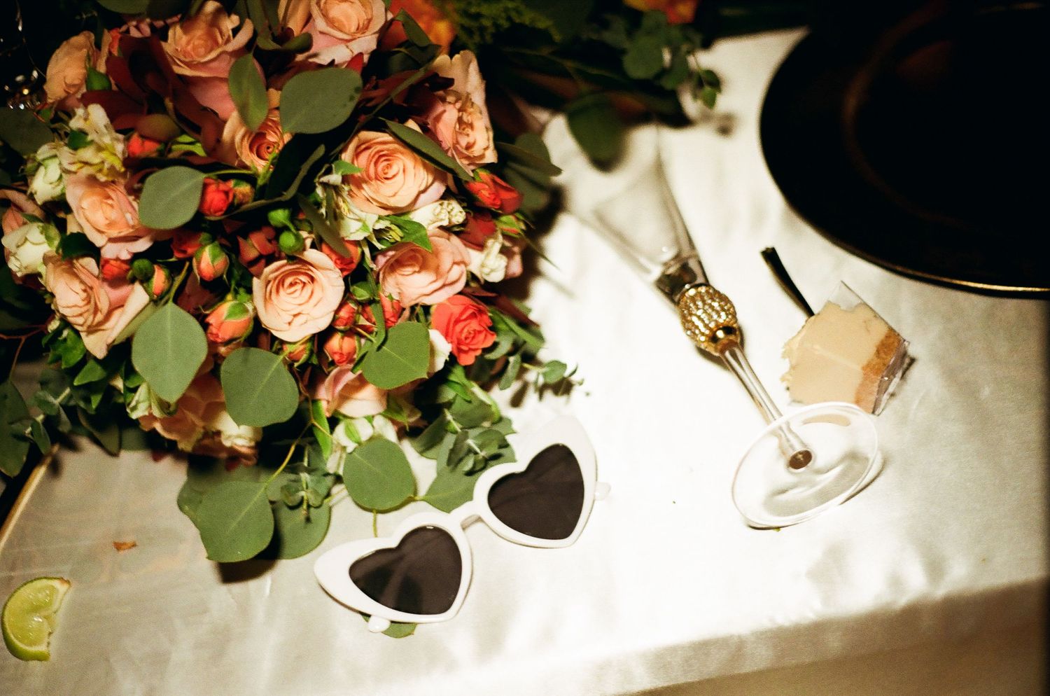 Wedding reception table details with heart-shaped sunglasses and coral roses on white linens.