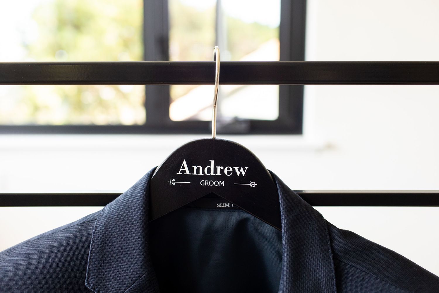 Black suit jacket hanging on a personalized wooden hanger against silhouetted window blinds.