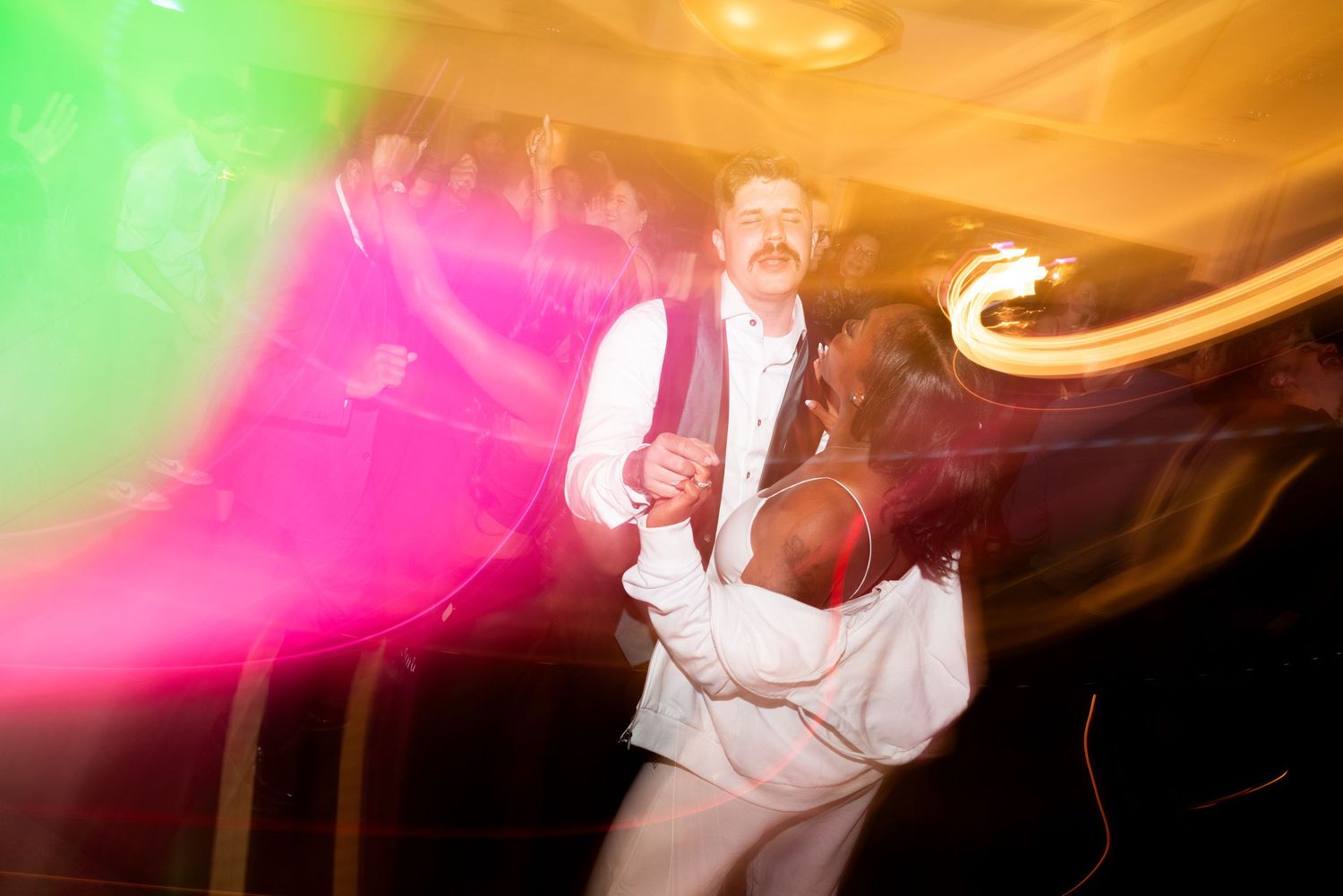 A couple dances beneath colorful pink and green party lights creating a vibrant motion blur effect.
