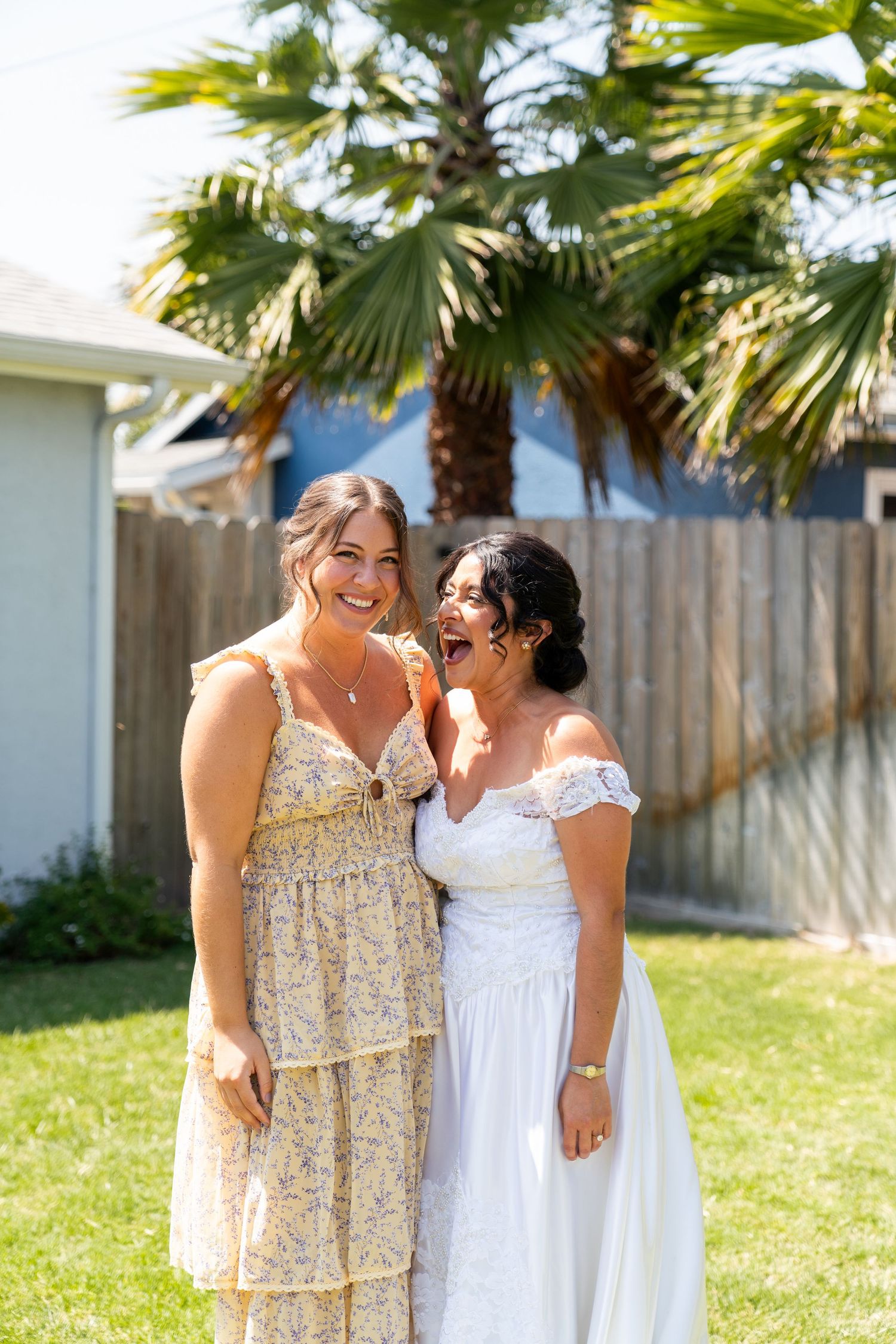 Two people wearing light-colored dresses share a joyful moment in a backyard with palm trees.