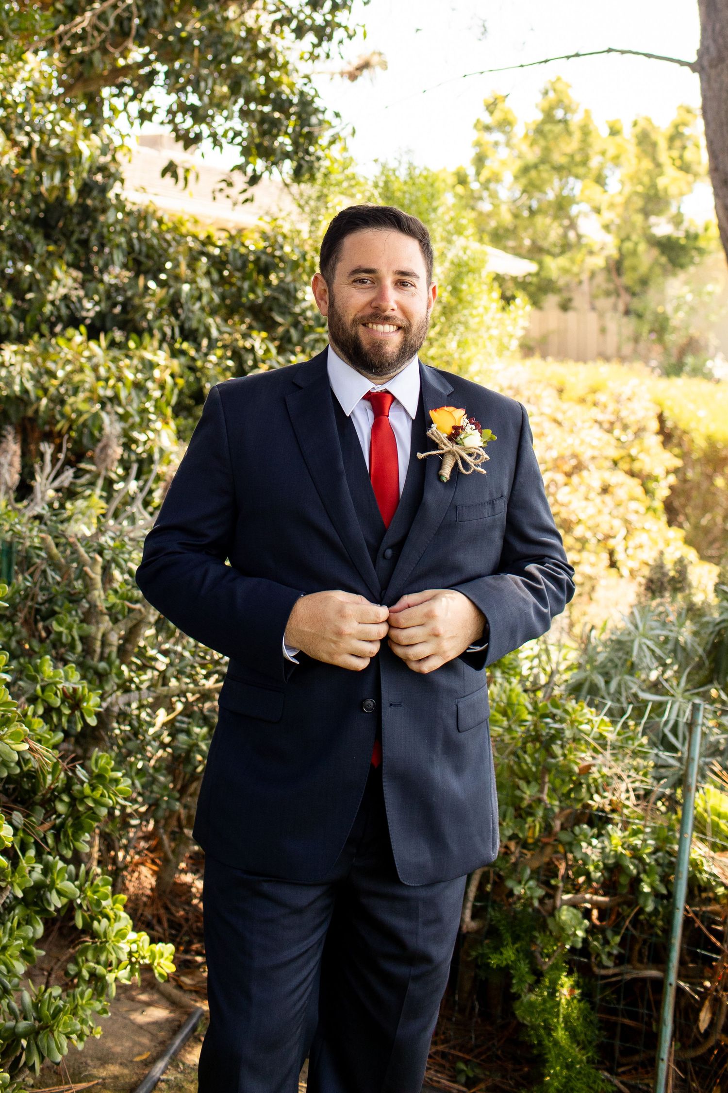 Groom in navy suit with red tie and boutonniere poses in garden setting.