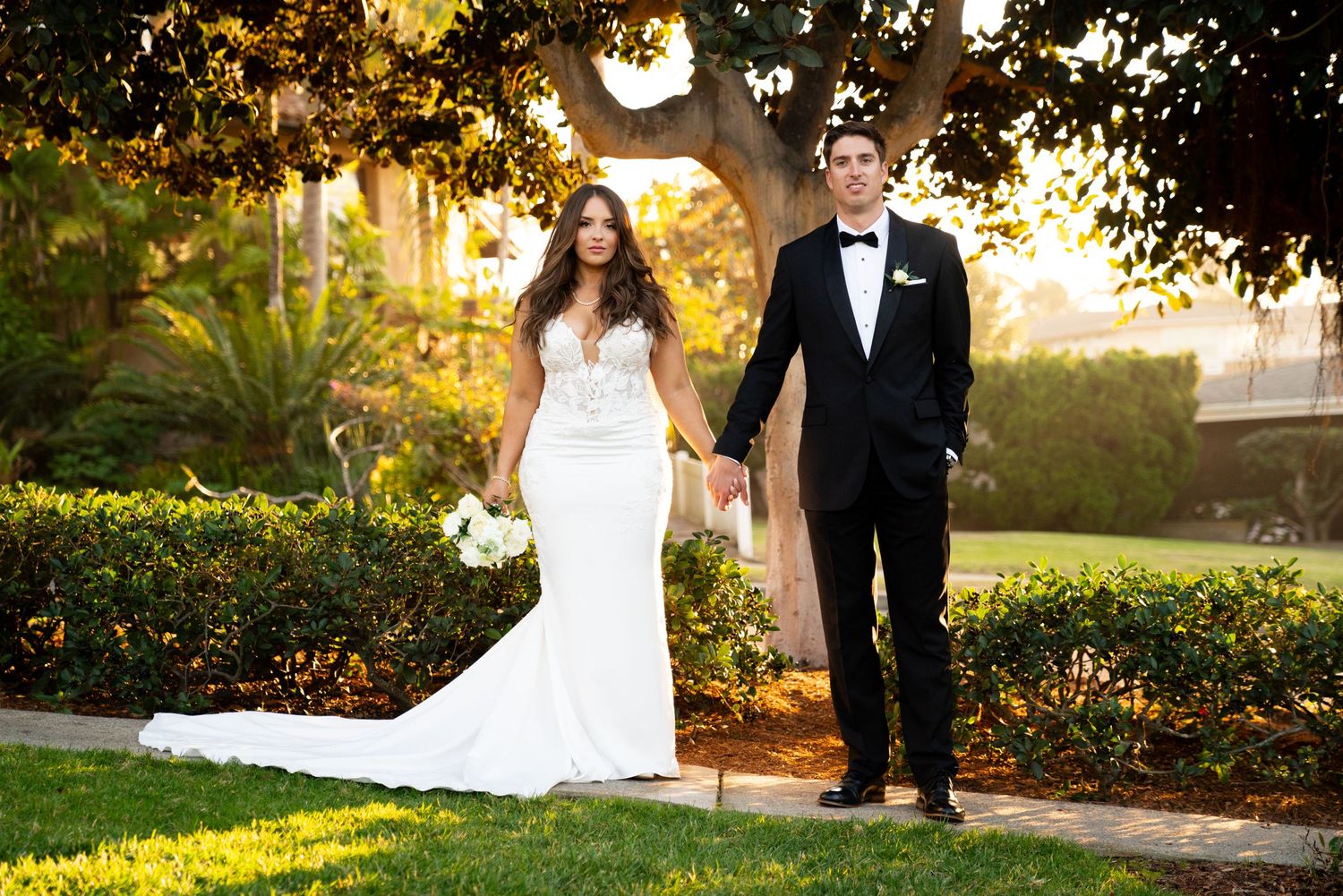 Newlyweds in formal attire holding hands while walking through garden archway at sunset.