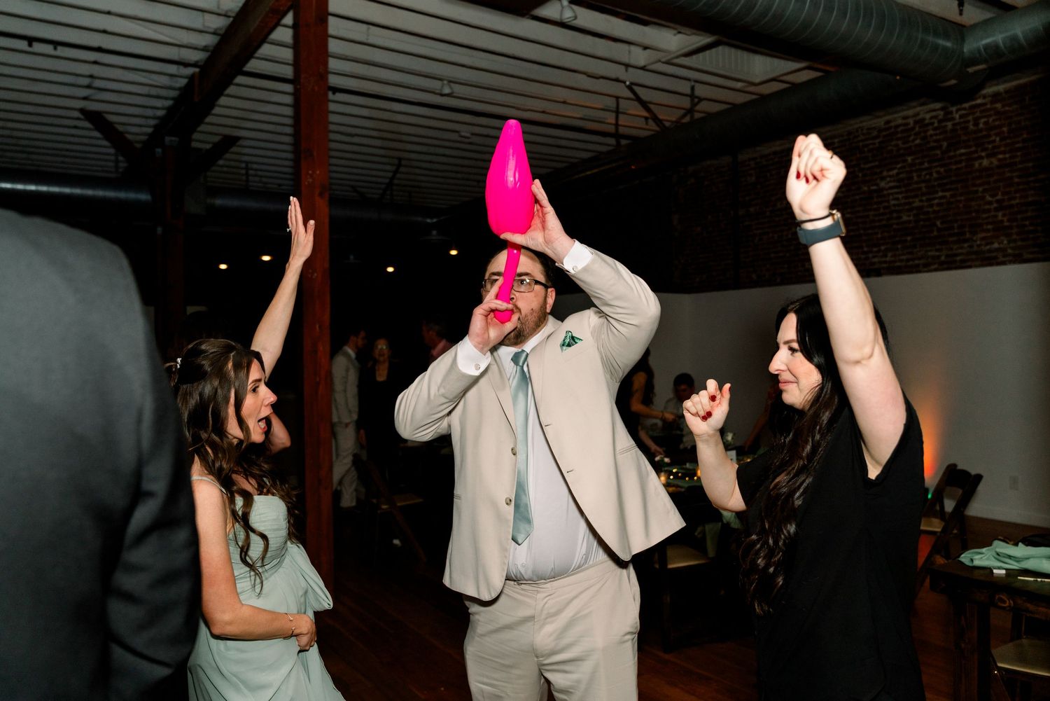 People dancing energetically at a party while someone in a gray suit holds up a bright pink object in celebration.
