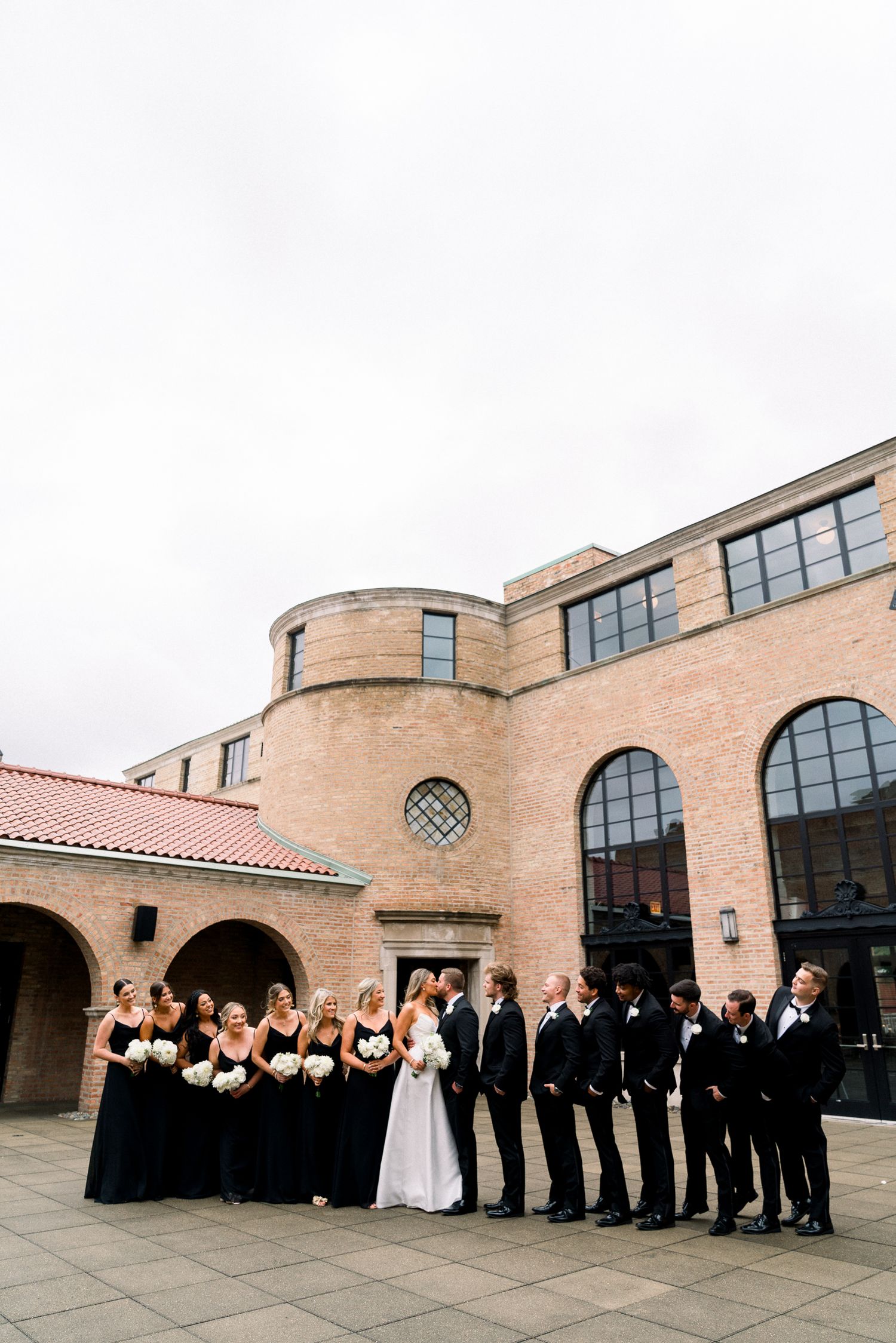 Wedding party in black and white formal attire lined up outside a Spanish-style building with arched windows and terracotta roof.