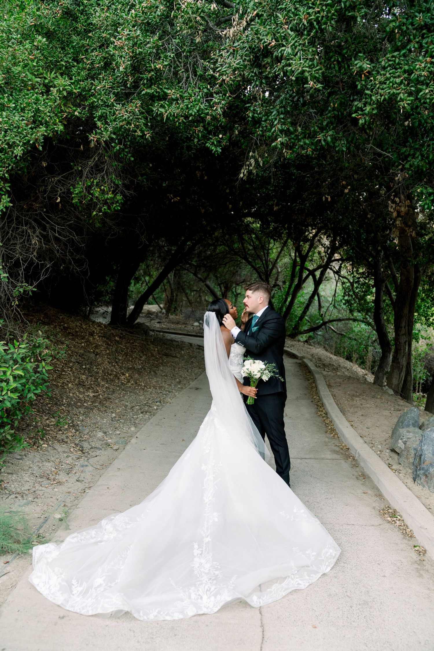 Bride in flowing white gown poses with groom on winding garden path under green trees.