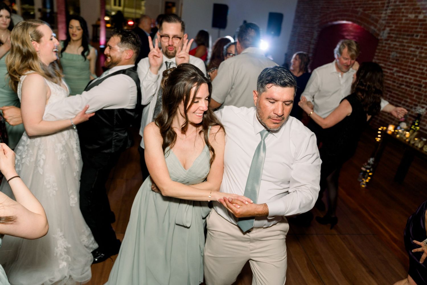 Party guests dancing together on a wood floor at a wedding reception with brick walls visible in the background.