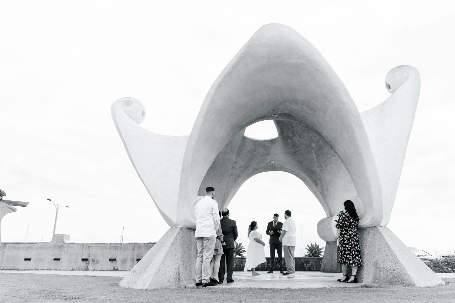 A black and white photograph of people gathered under a large modernist concrete arch structure.
