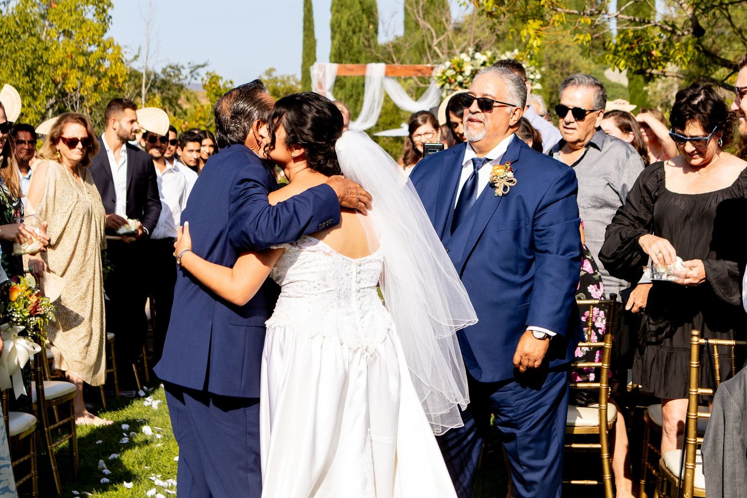 An outdoor wedding ceremony with guests watching under bright sunlight and autumn foliage.